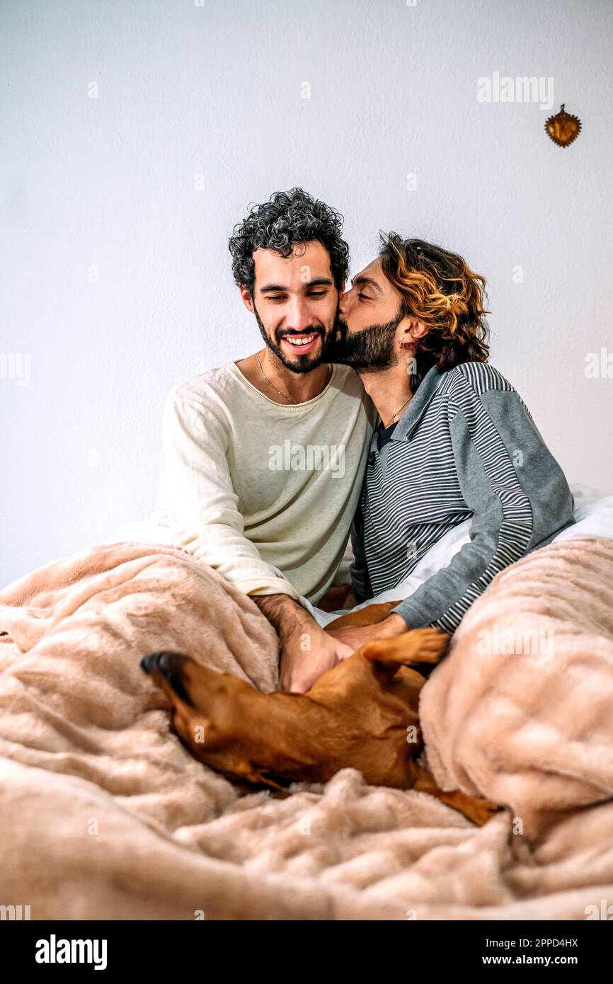 Schwuler Mann, der seinen Freund küsst und zu Hause mit dem Hund im Bett spielt Stockfoto
