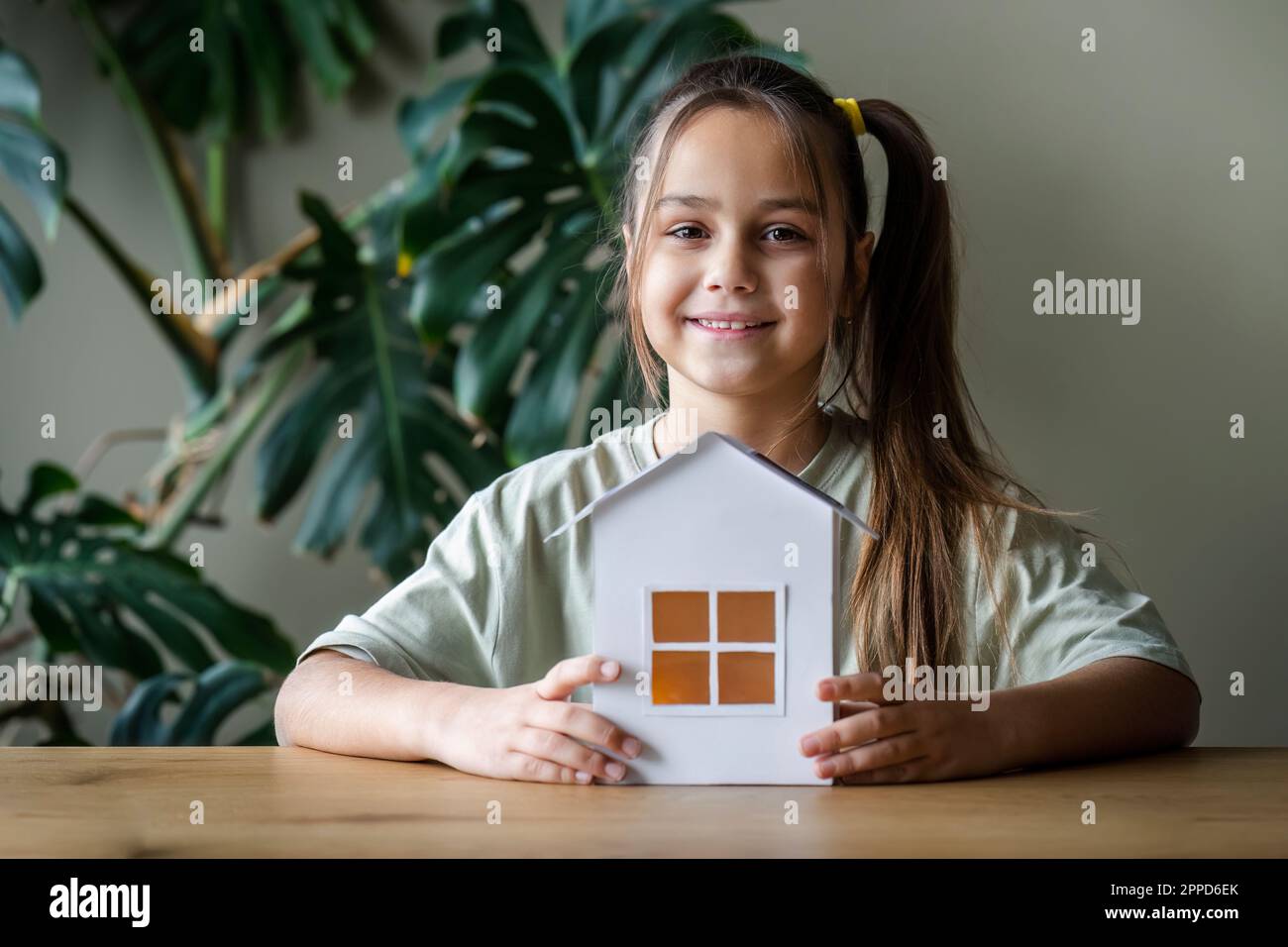 Lächelndes Mädchen sitzt mit grünem Hausmodell am Tisch Stockfoto