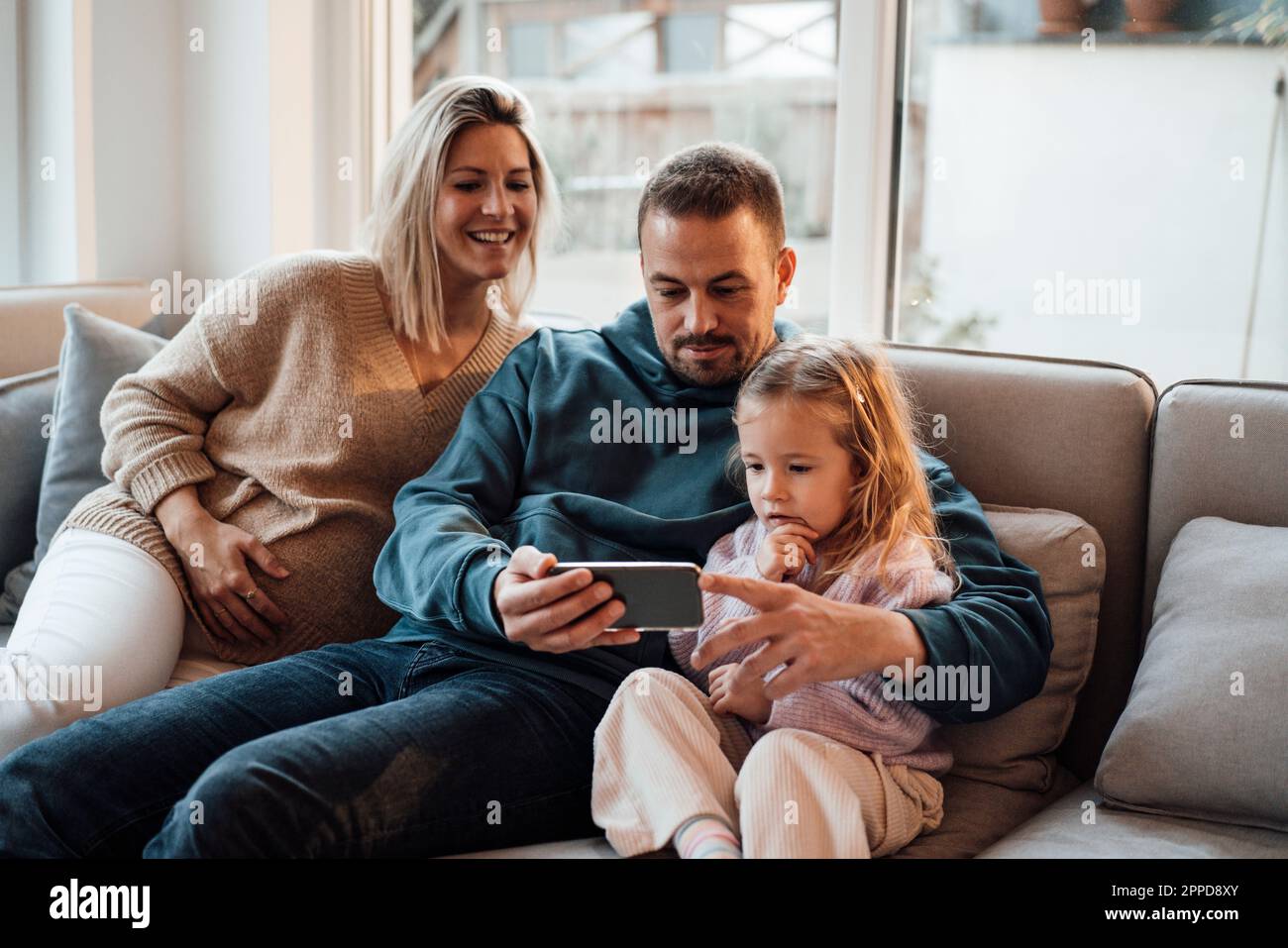 Eltern teilen sich ihr Smartphone mit ihrer Tochter, die zu Hause auf dem Sofa sitzt Stockfoto