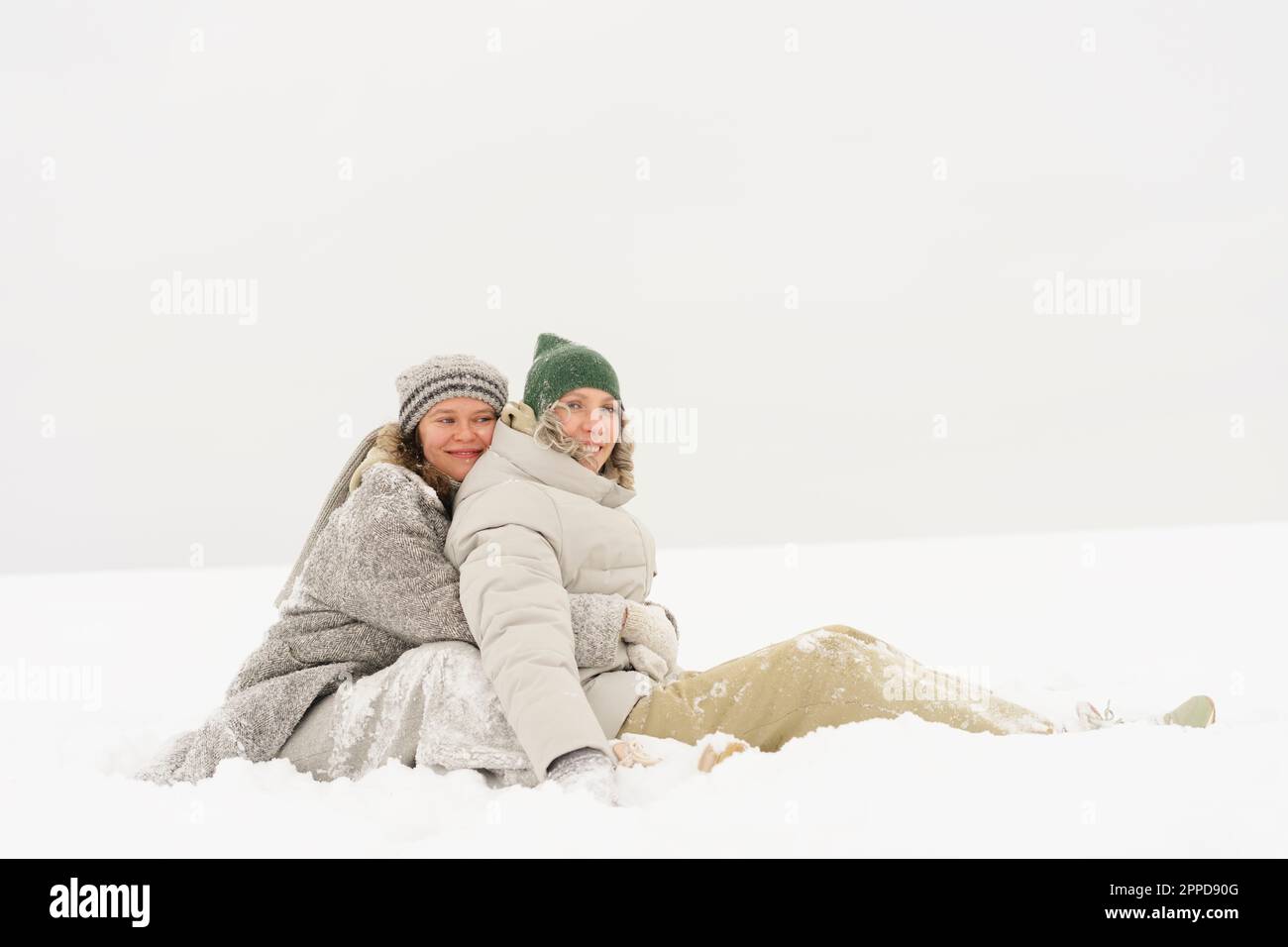 Eine Frau umarmt eine Freundin von hinten, sitzt im Schnee Stockfoto