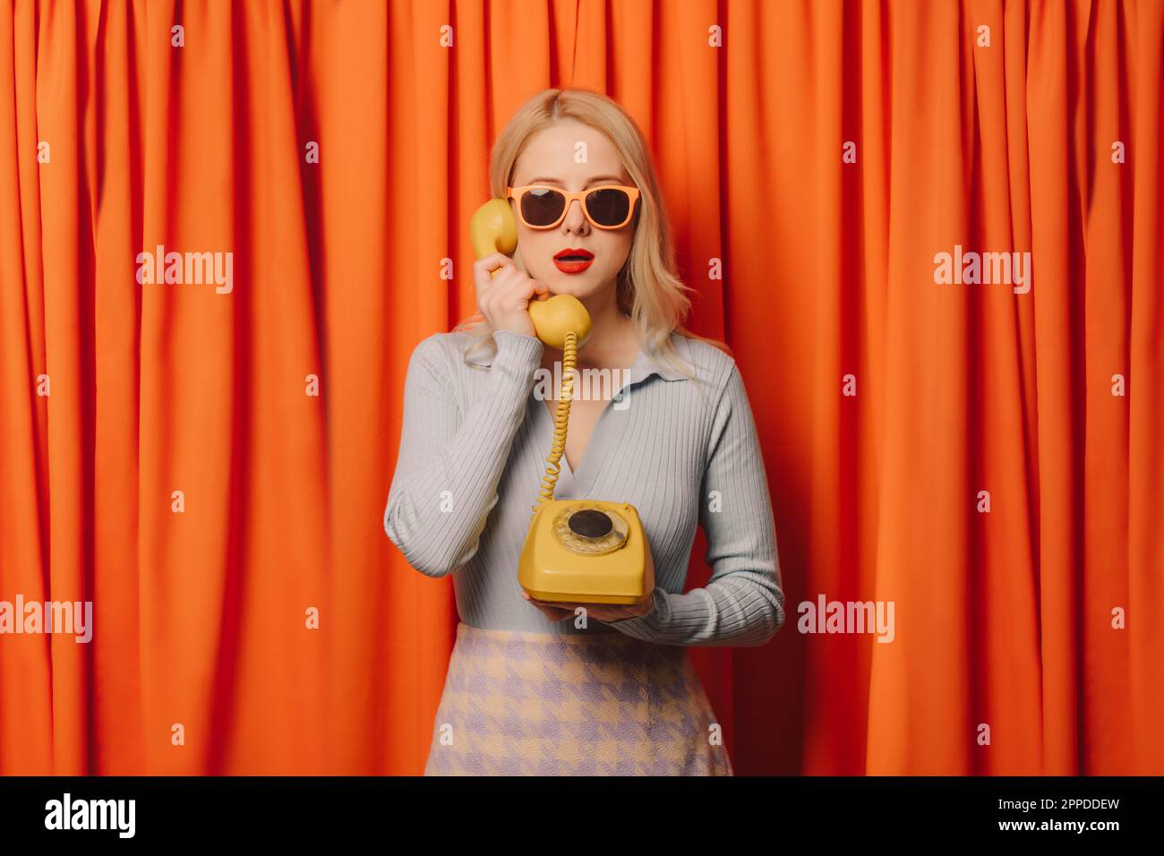 Eine Frau, die vor orangefarbenen Vorhängen auf einem alten Festnetztelefon spricht Stockfoto Eine Frau, die vor orangefarbenen Vorhängen auf einem alten Festnetztelefon spricht Stockfoto