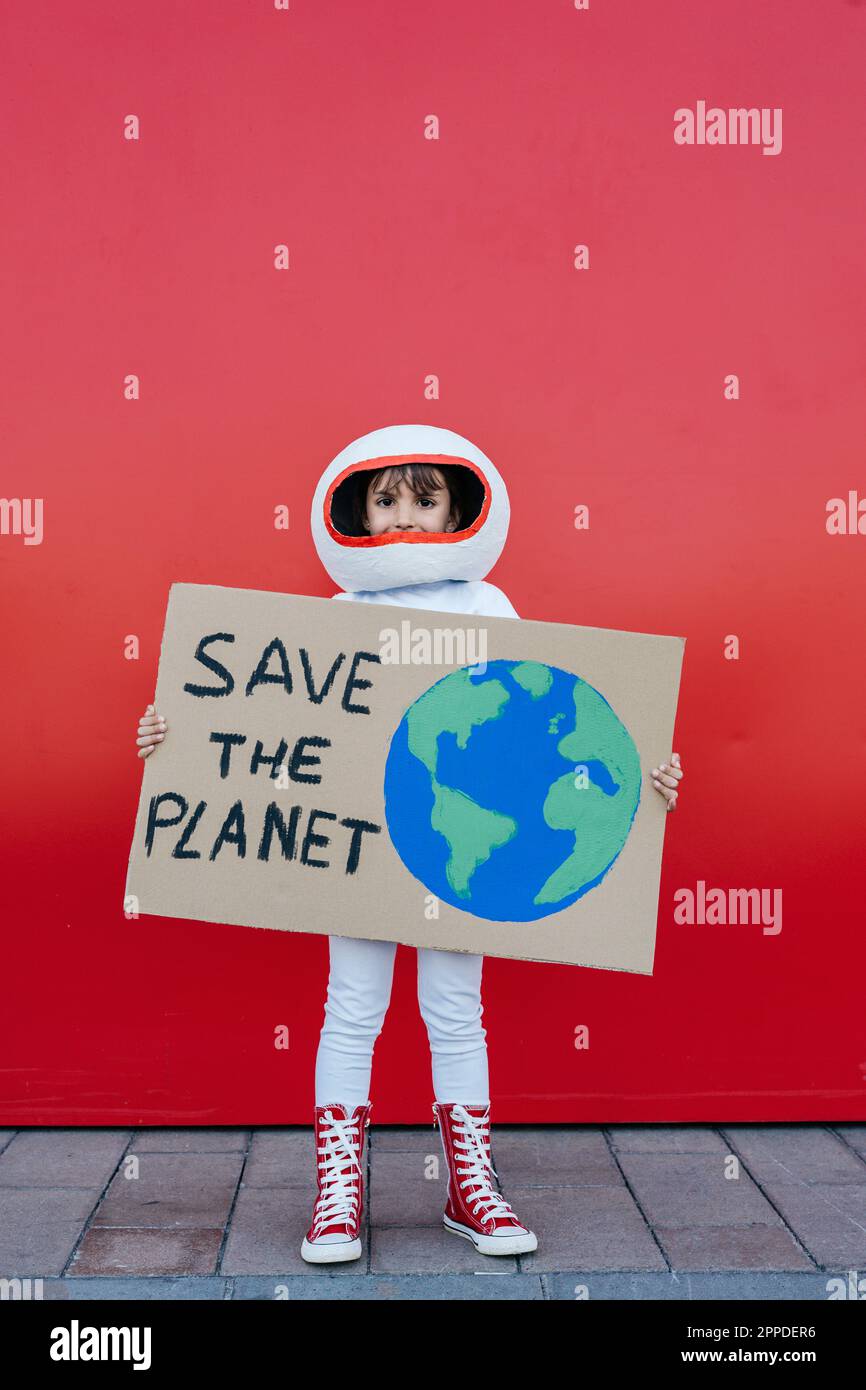 Ein Mädchen in Astronautenkostüm, das vor der roten Wand steht und die Plakette „Save the Planet“ hält Stockfoto