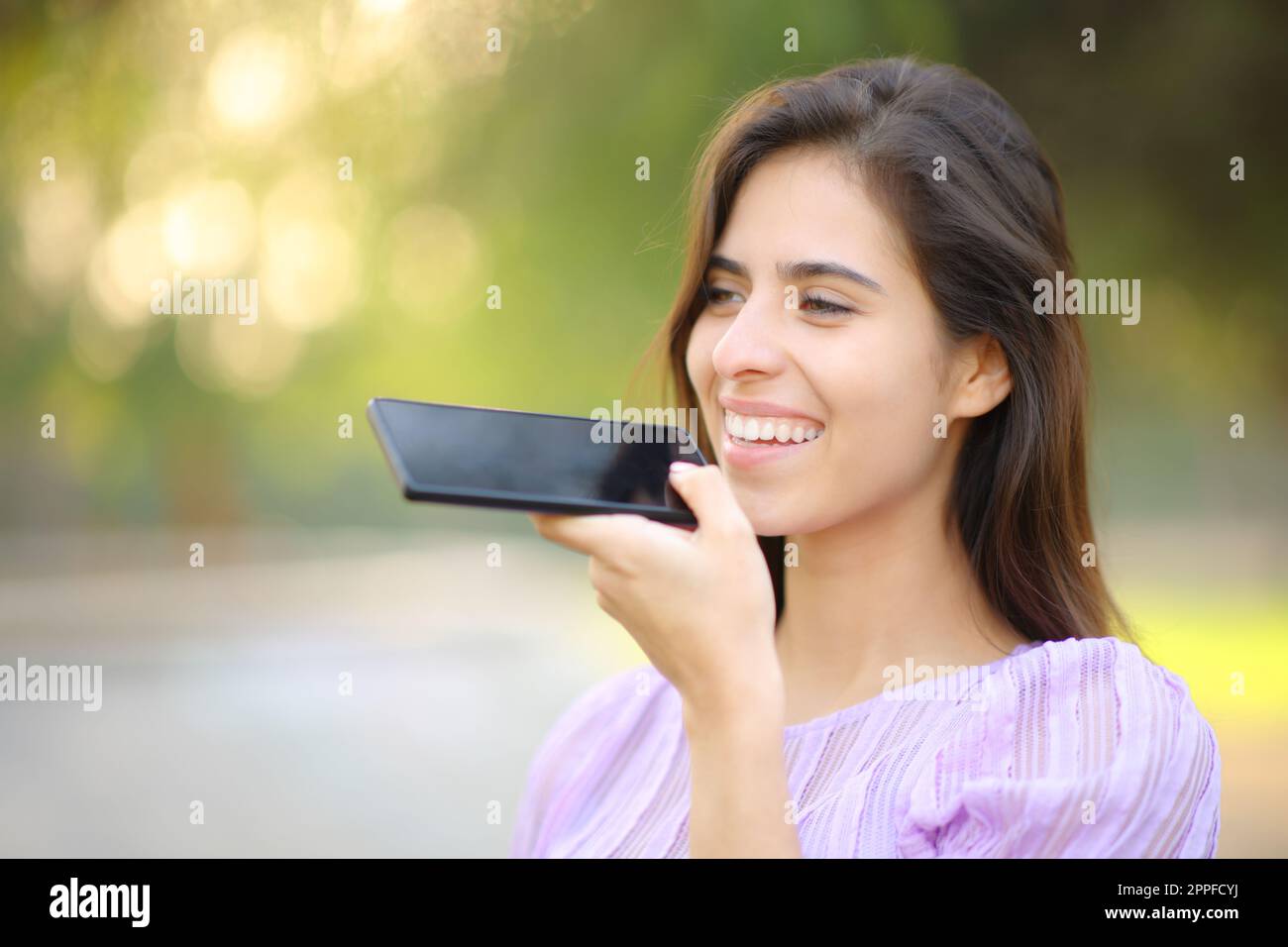 Glückliche Frau, die in einem Park Spracherkennung am Handy benutzt Stockfoto