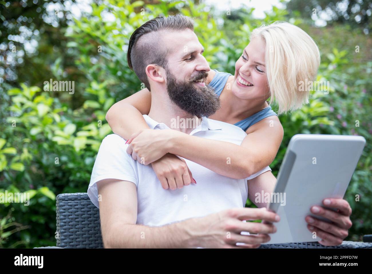 Junger Mann, der ein digitales Tablet benutzt, mit seiner Freundin, die sich von hinten umarmt, in Garten, Bayern, Deutschland Stockfoto