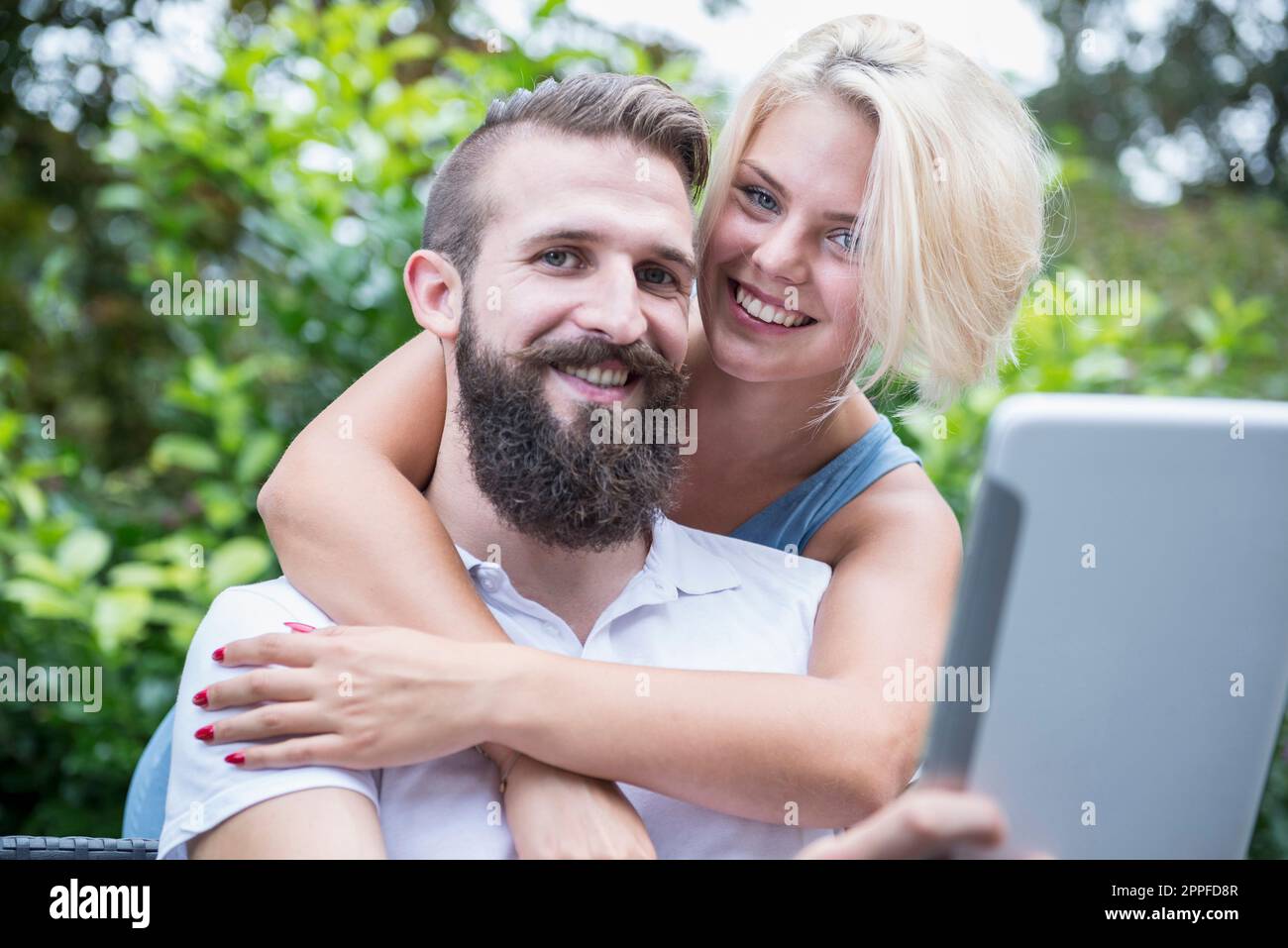 Junger Mann, der ein digitales Tablet benutzt, mit seiner Freundin, die sich von hinten umarmt, in Garten, Bayern, Deutschland Stockfoto