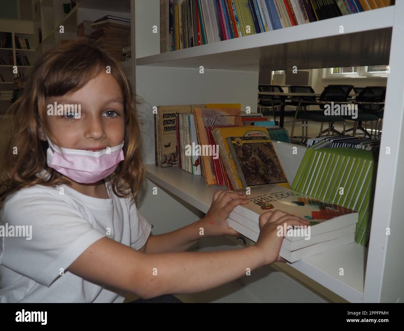 Sremska Mitrovica, Serbien, 15. Mai 2021. Ein 7-jähriges Mädchen, das eine Maske und ein weißes T-Shirt trägt, steht neben einem Bücherregal in einer Bibliothek und berührt Bücher mit Händen. Schulbildung, Literatur und Lesen Stockfoto