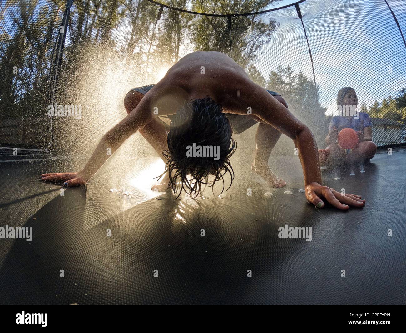 Ein Junge, der sich bei Sonnenuntergang mit einem Sprinkler auf Trampolin bückt Stockfoto