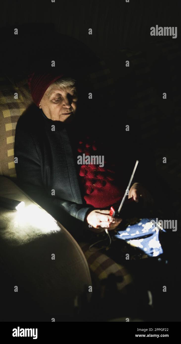 Ukraine: Großmutter hält während russischer Terroranschläge Radio in den Händen, während sie im Dunkeln des Blackouts sitzt. Seniorin in Winterkleidung in Dunkelheit auf dem Sofa mit Handfunkempfänger, der Nachrichten hört Stockfoto