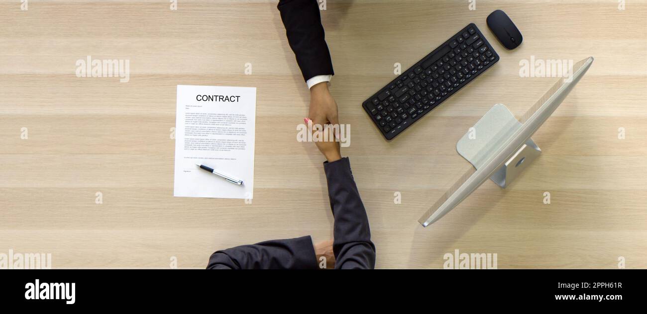 Der asiatische Human Resource Manager in schwarzem Anzug schüttelt sich die Hände, um dem jungen Kandidaten nach dem Interview zu gratulieren. Computermonitor, Tastatur, Maus und Vertragsdokument sind auf Holztisch. Draufsicht Stockfoto