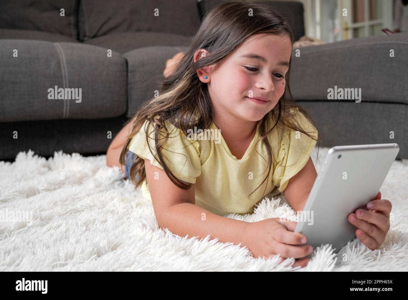 Ein süßes kleines Mädchen sieht sich Cartoons auf einem digitalen Tablet an. Das Kind liegt auf dem Boden und lacht mit einem elektronischen Gerät. Freizeit für Kinder im Haus. Stockfoto