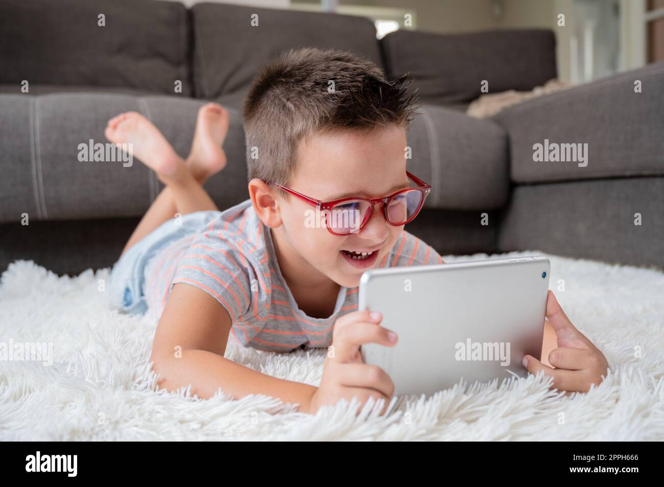 Süßer kleiner Junge, schau dir Cartoons auf einem digitalen Tablet an. Das Kind liegt auf dem Boden und lacht mit einem elektronischen Gerät. Freizeit für Kinder im Haus. Stockfoto