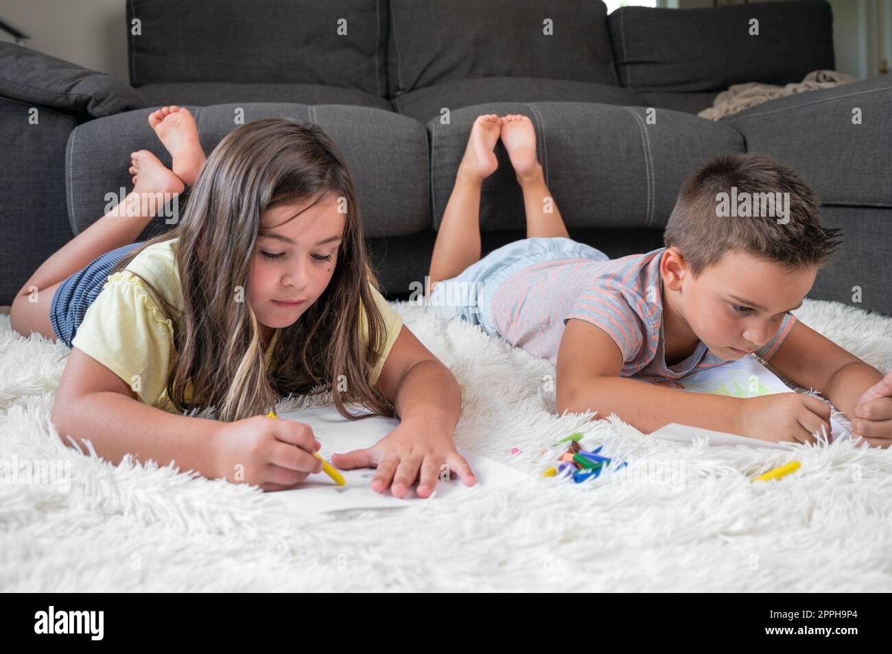 Geschwister, die zu Hause zusammen spielen. Ein kleiner Junge und ein Mädchen, die auf dem Teppich liegen und auf weißen Blättern mit bunten Buntstiften zeichnen Stockfoto