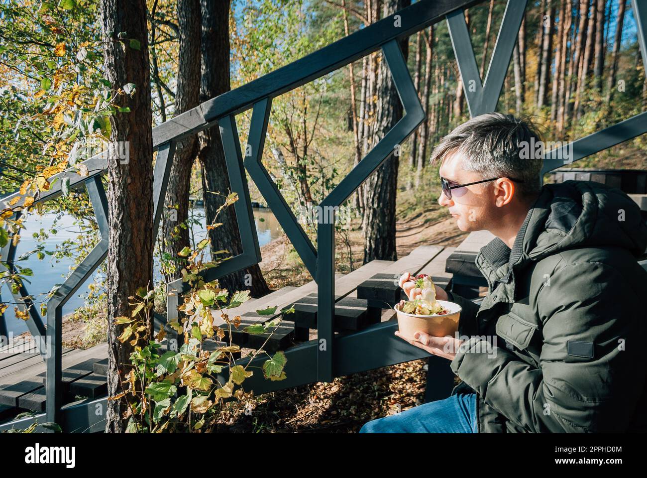 Ein Mann sitzt am Ufer des Baltieji Lakajai im Labanoras Regional Park, Litauen, in einer warmen Kurta und isst einen Salat aus Einwegpapiergerichten, während er sich entspannt. Stockfoto