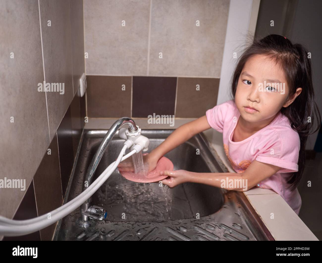 Ein bezauberndes Mädchen, das Geschirr in der Hausküche spült. Das Kind hat Spaß dabei, seinen Eltern bei der Hausarbeit zu helfen. Stockfoto