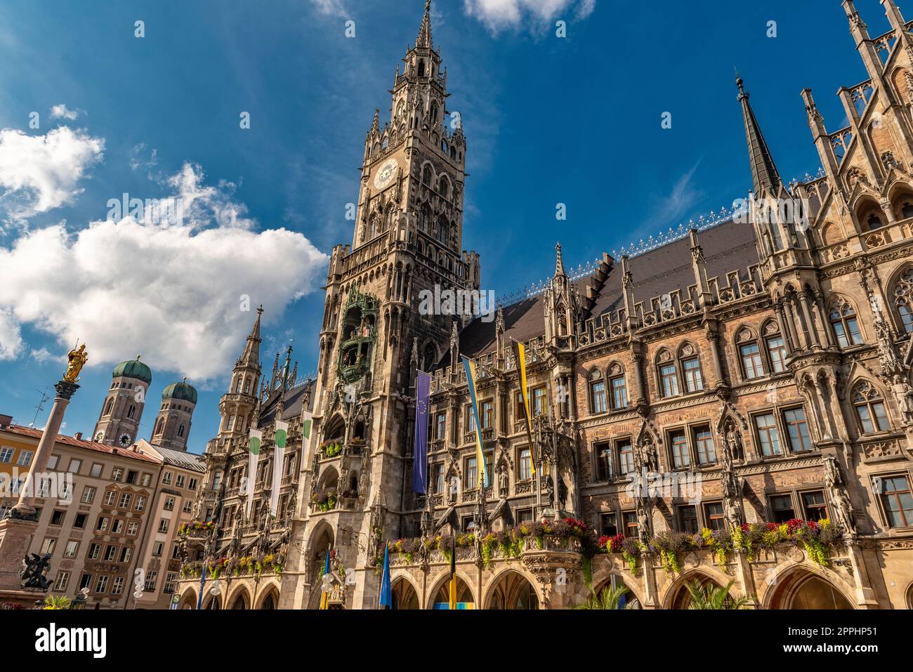 Die neogotische Fassade des neuen Rathauses mit Rathausturm in der Münchner Altstadt im Blick von unten vom Marienplatz bei schönem Sommerwetter und blauem Himmel mit den beiden Türmen der Frauenkirche im Hintergrund Stockfoto