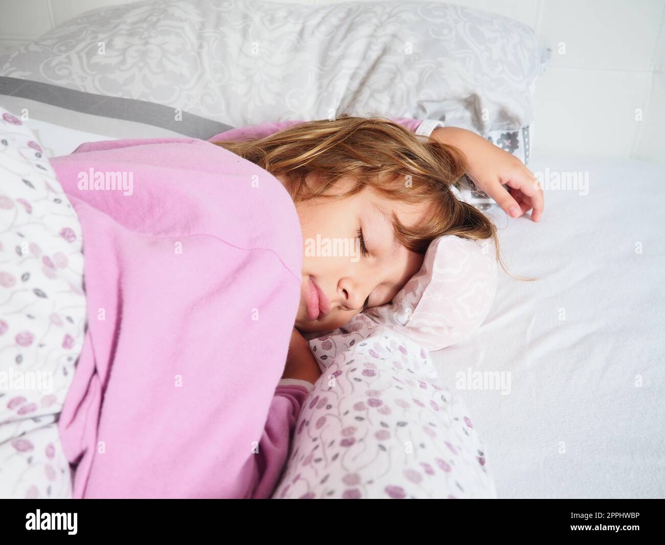 Ein hübsches, 8 Jahre altes, weißes Mädchen mit blonden Haaren, gekleidet in pinkfarbenen Pyjamas, schläft auf einem Bett mit einer flauschigen Decke, umarmt ein Kissen. Sanftes Licht der Morgensonne strömt durch das Fenster. Stockfoto