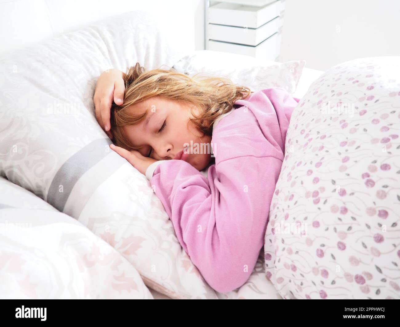 Ein hübsches, 8 Jahre altes, weißes Mädchen mit blonden Haaren, gekleidet in pinkfarbenen Pyjamas, schläft auf einem Bett mit einer flauschigen Decke, umarmt ein Kissen. Sanftes Licht der Morgensonne strömt durch das Fenster. Stockfoto