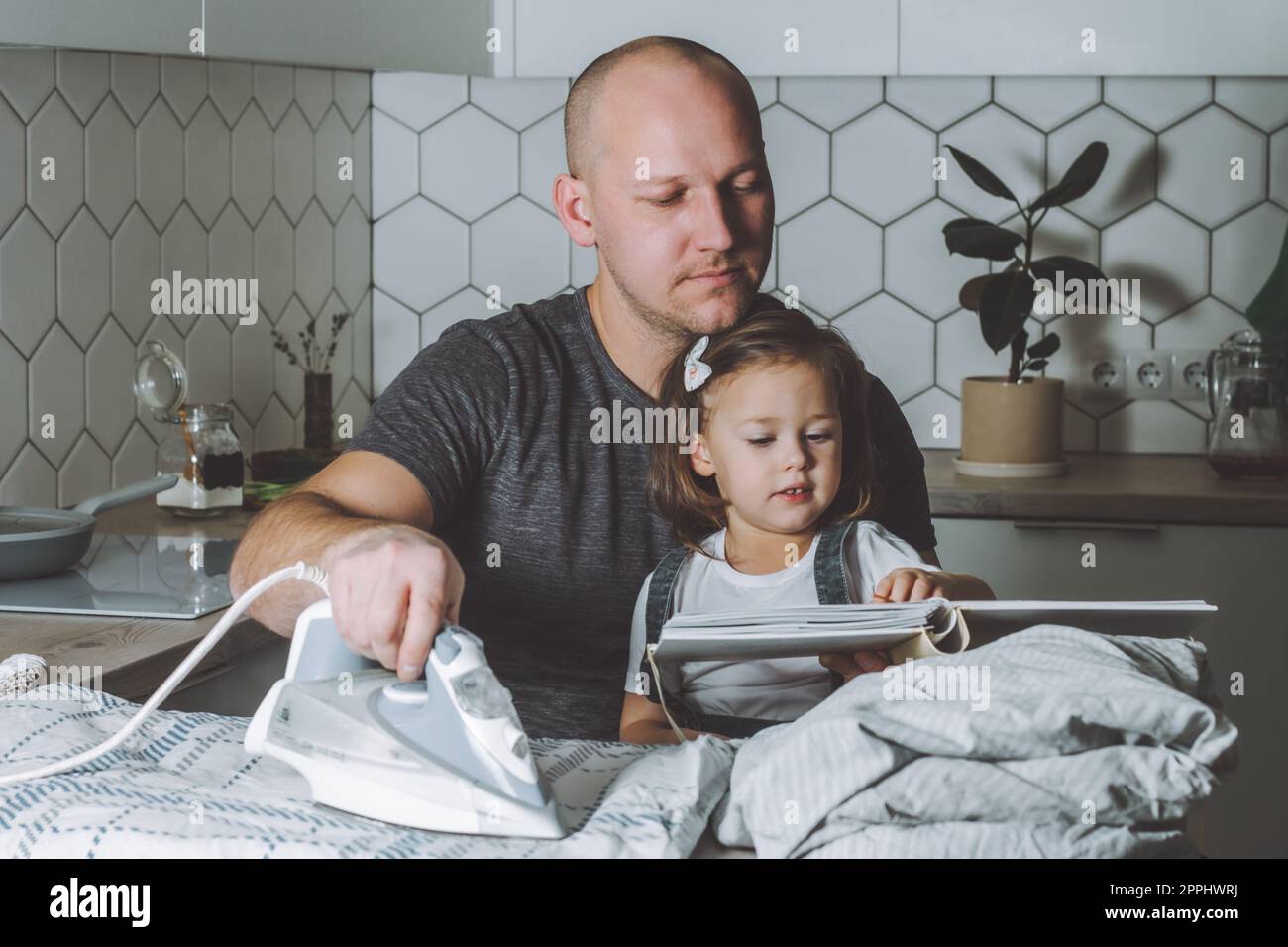 Mann bügeln Bettwäsche und Buch zu lesen, um seine kleine Tochter. Vater ist in Hausarbeit beschäftigt. Stockfoto