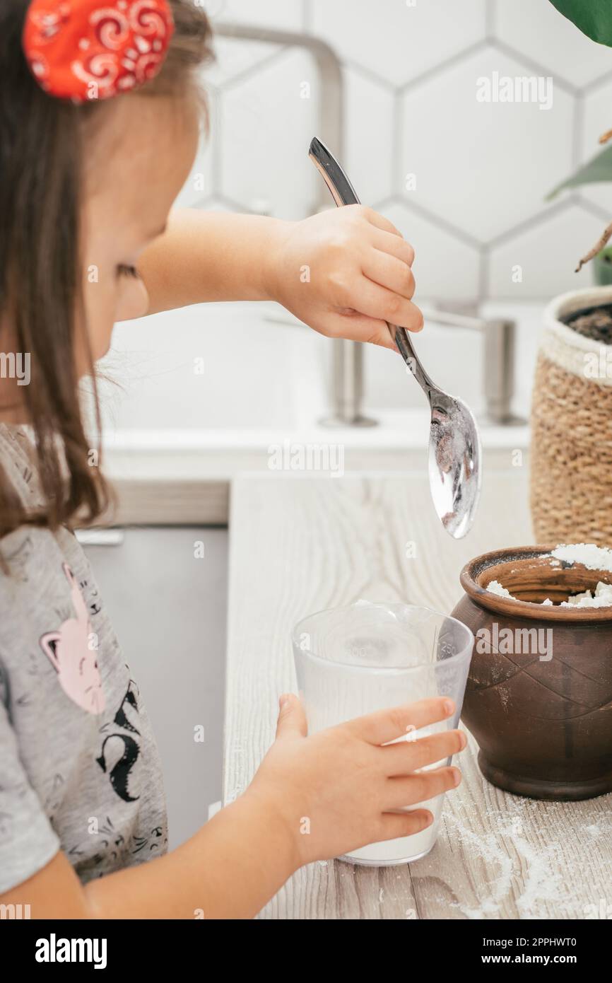 Ein dunkelhaariges Mädchen, 3 Jahre alt, mit Haarband, backt Apfelkuchen in der Küche. Das Kind gibt Mehl in den Messbecher. Vertikal Stockfoto