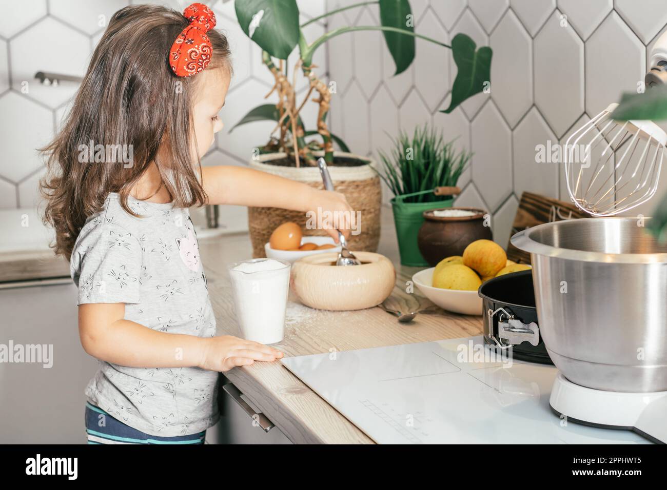 Ein dunkelhaariges Mädchen, 3 Jahre alt, mit rotem Haarband, backt Apfelkuchen in der Küche. Das Kind gibt Zucker in den Messbecher Stockfoto
