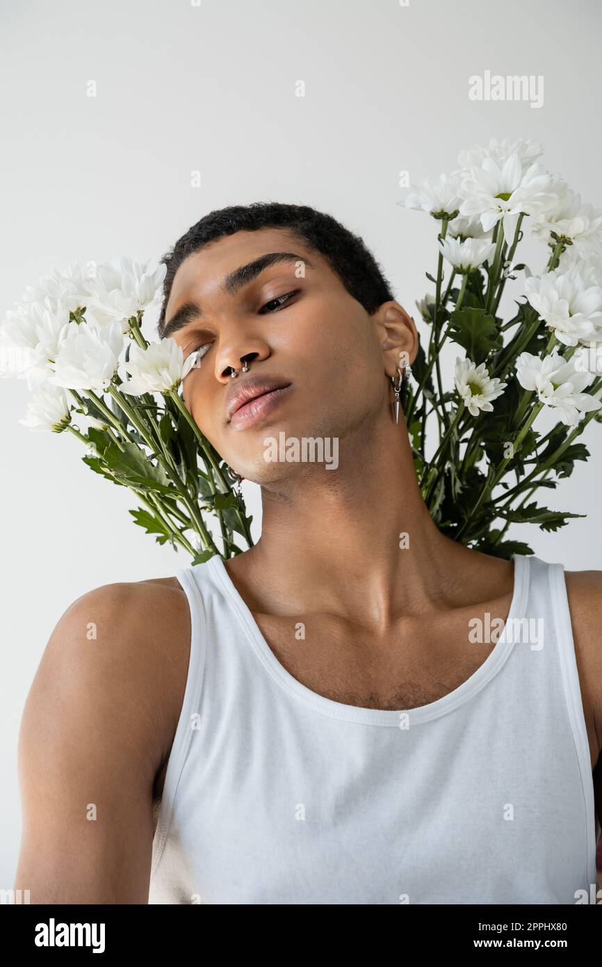 Porträt eines jungen afroamerikanischen Kerls in weißem Panzeroberteil mit weißen Chrysanthemen isoliert auf grauem Stockbild Stockfoto