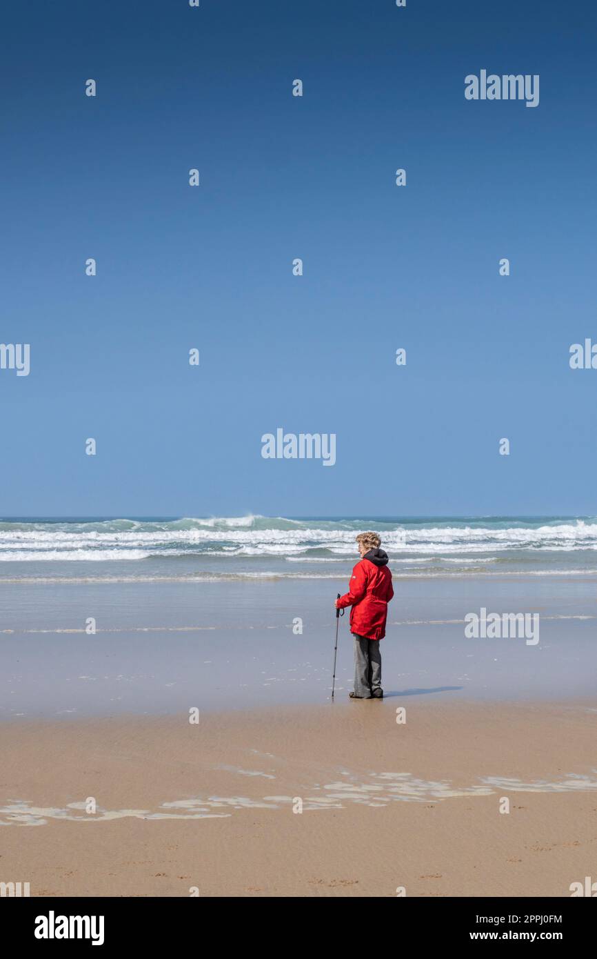 Eine reife Frau, die einen leuchtend roten Mantel trägt und allein am Strand von Mawgan Porth in Cornwall in Großbritannien steht. Stockfoto