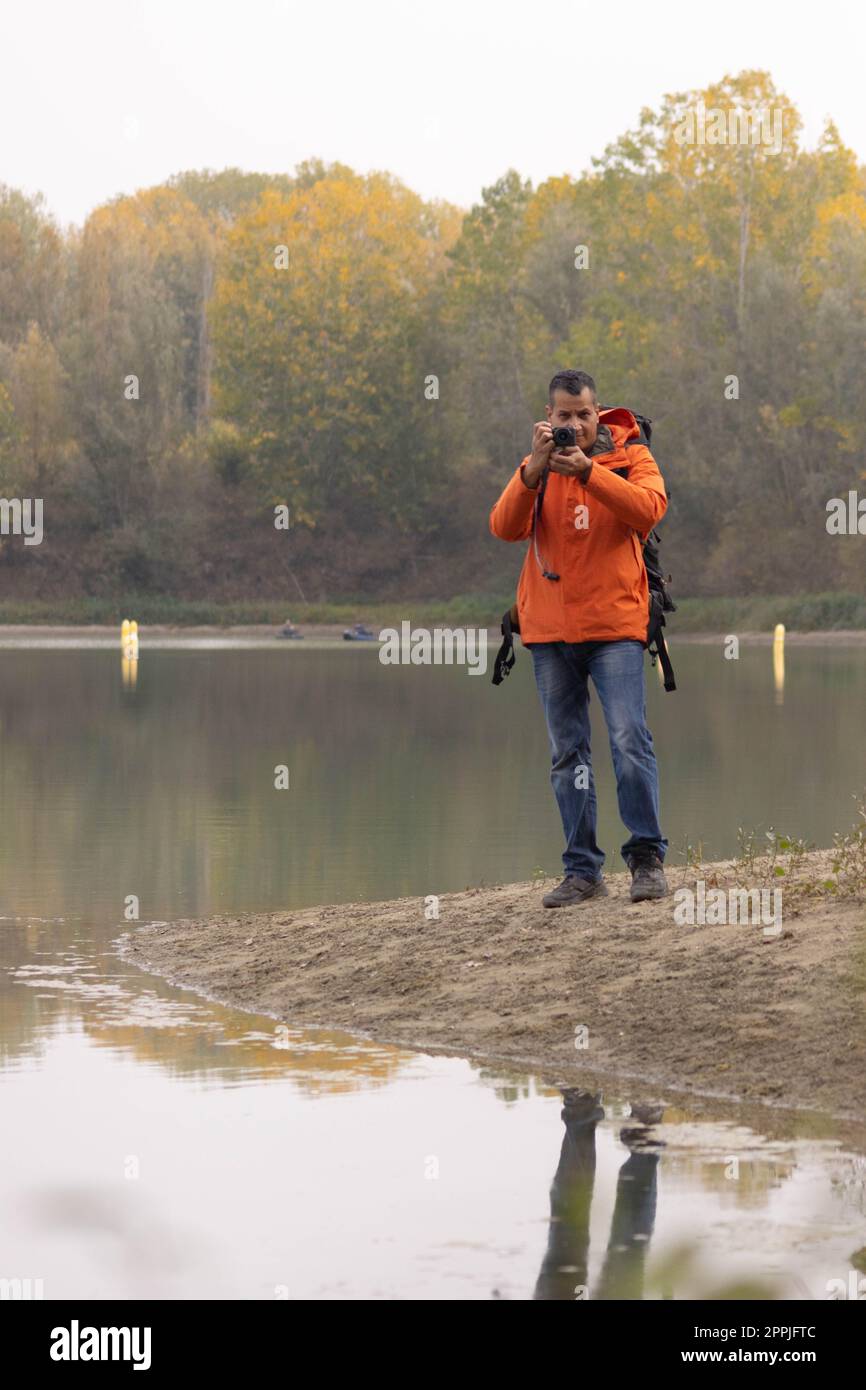 Porträt eines Reisenden am See im Herbst, der ein Foto mit der Digitalkamera und der orangefarbenen Jacke machte Stockfoto