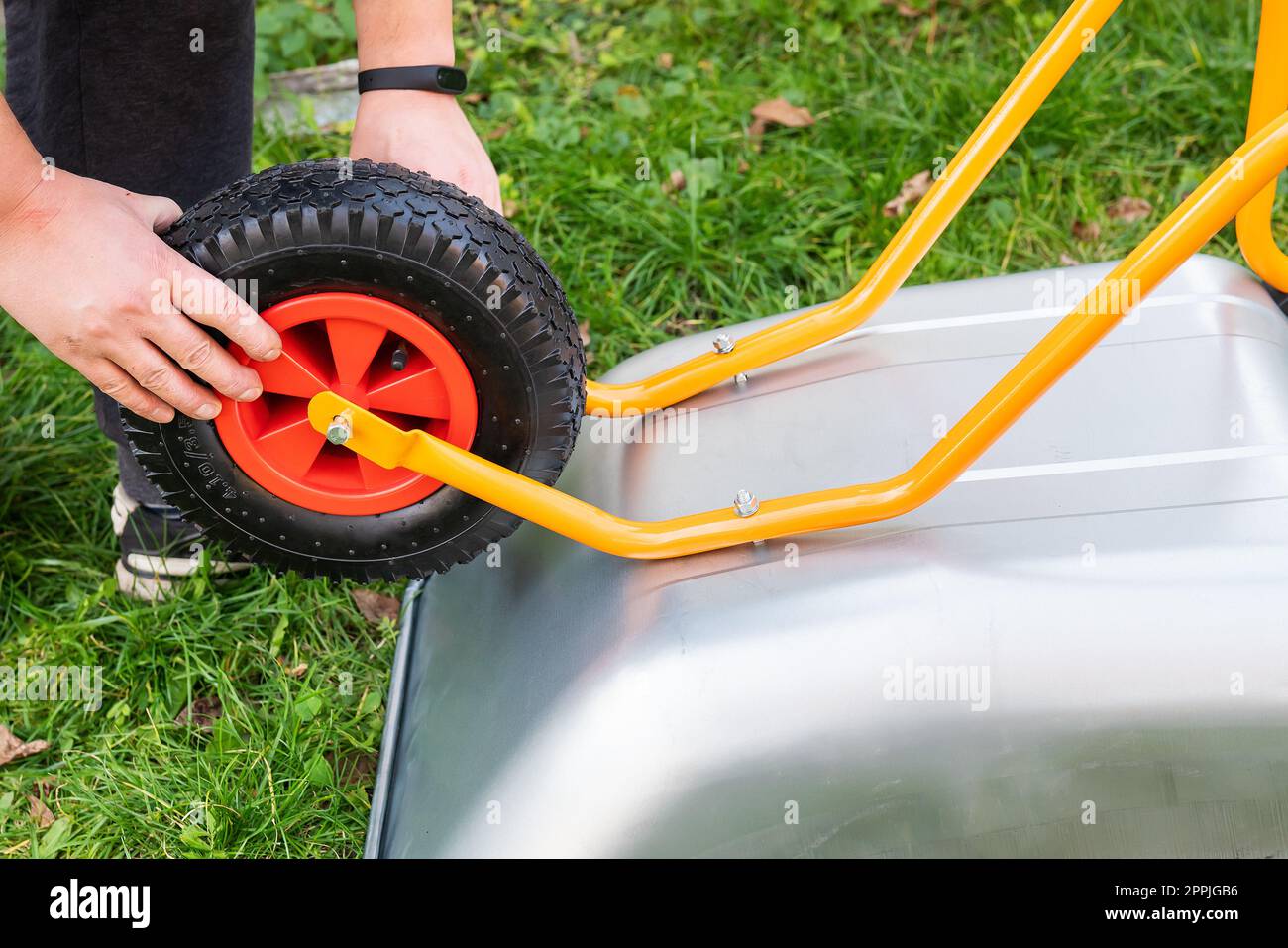 Auf der Straße ist eine Schubkarre aus Metall, ein Mann dreht eine neue Schubkarre für die Arbeit, zieht Schrauben und Muttern fest, hängt ein Rad auf. Stockfoto