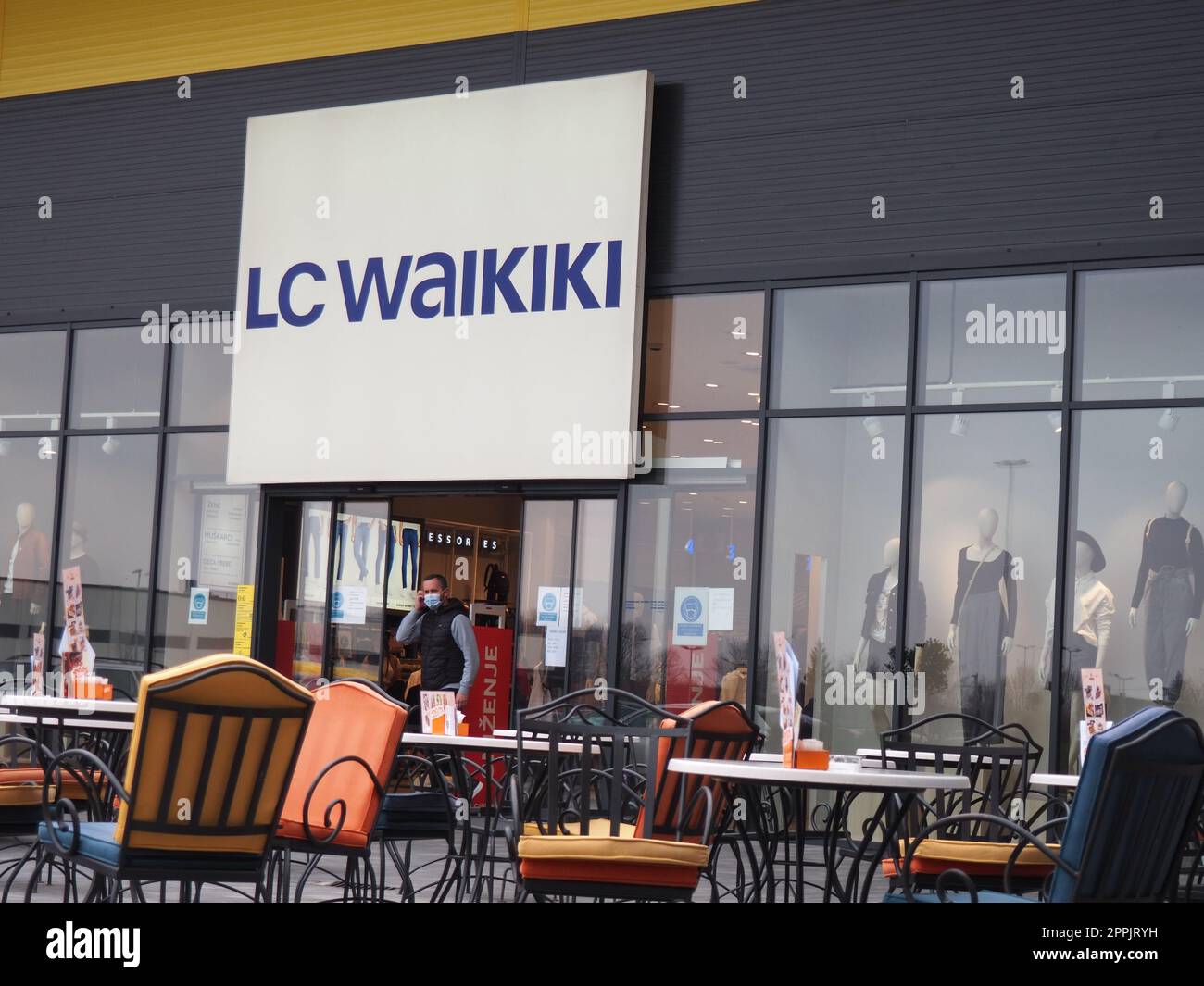 Sremska Mitrovica, Serbien, 27. Februar 2021 Einkaufszentrum, Bekleidungsgeschäft LC WAIKIKI. Ein Mann, der eine chirurgische Schutzmaske trägt, geht aus den Schiebetüren eines Ladens. Café mit Stühlen und Tischen aus Metall Stockfoto