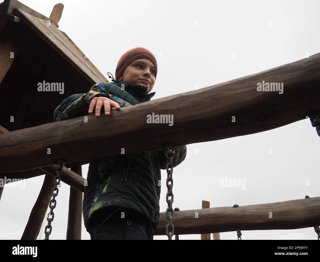 Ein 8-jähriger Junge auf dem Spielplatz. Das Kind trägt warme Kleidung für die Demi-Saison - einen braunen Hut und eine grüne Jacke. Kletterbereich aus Holz für Kinder. Ein Junge, der wegschaut Stockfoto