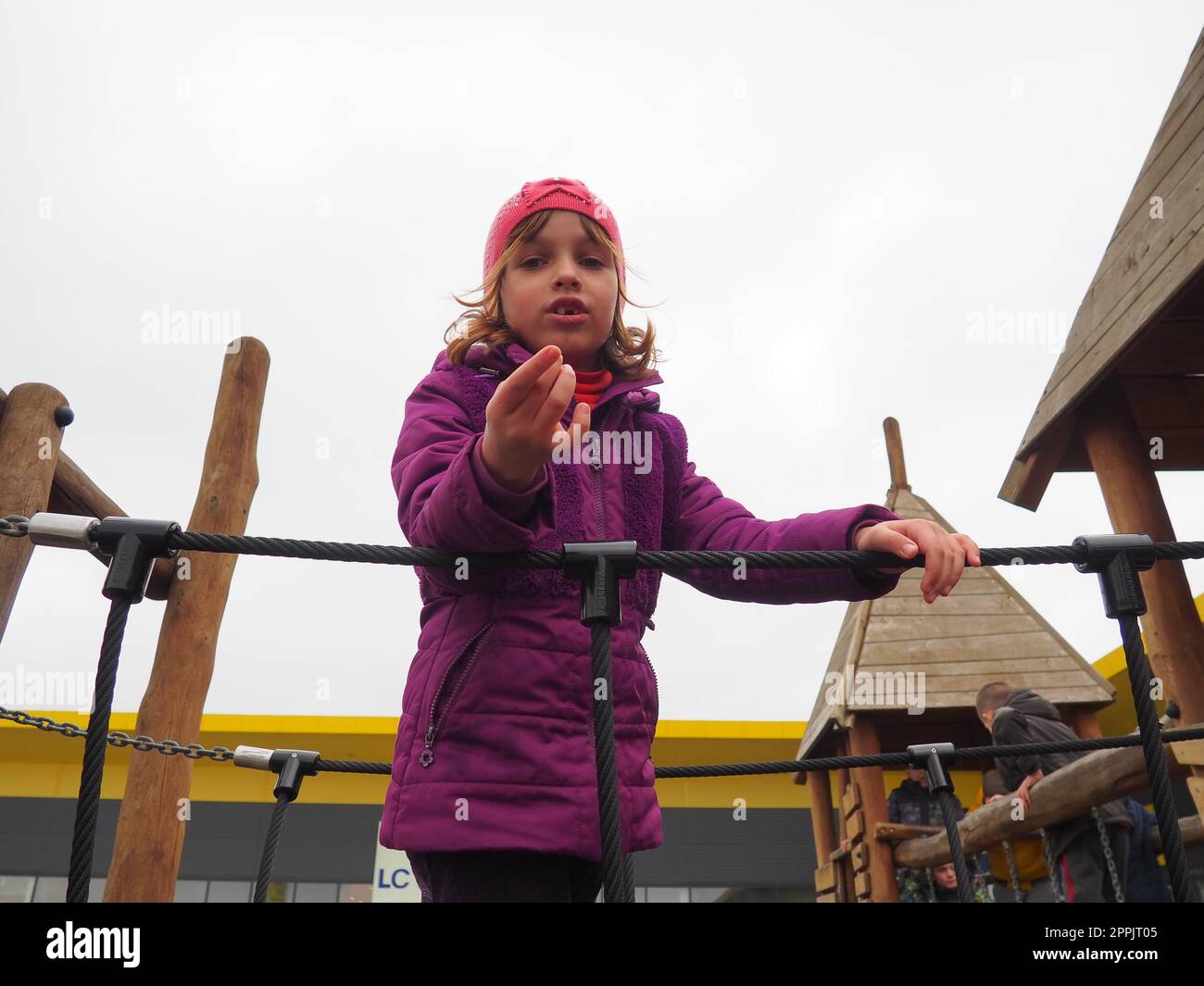 Mädchen 7 Jahre alt auf dem Spielplatz. Das Kind trägt warme Kleidung für die Halbzeit - eine lila Jacke und eine pinkfarbene Mütze. Kletterspielplatz für Kinder. Das Mädchen streckt ihre rechte Hand nach vorne aus Stockfoto