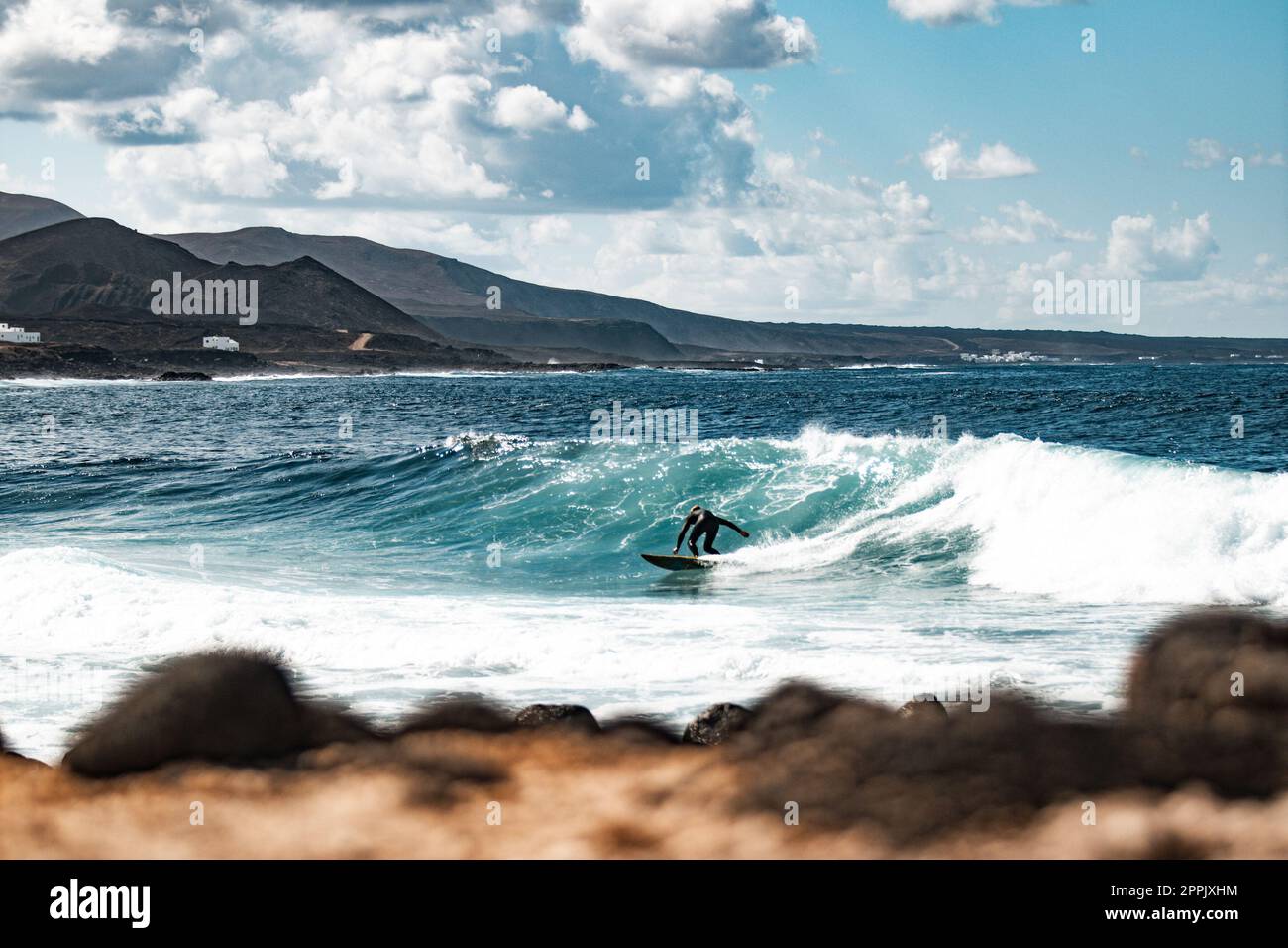 Wilde felsige Küste des Surfspots La Santa Lanzarote, Kanarische Inseln, Spanien. Surfer reiten eine große Welle in felsiger Bucht, Vulkan Berg im Hintergrund. Stockfoto