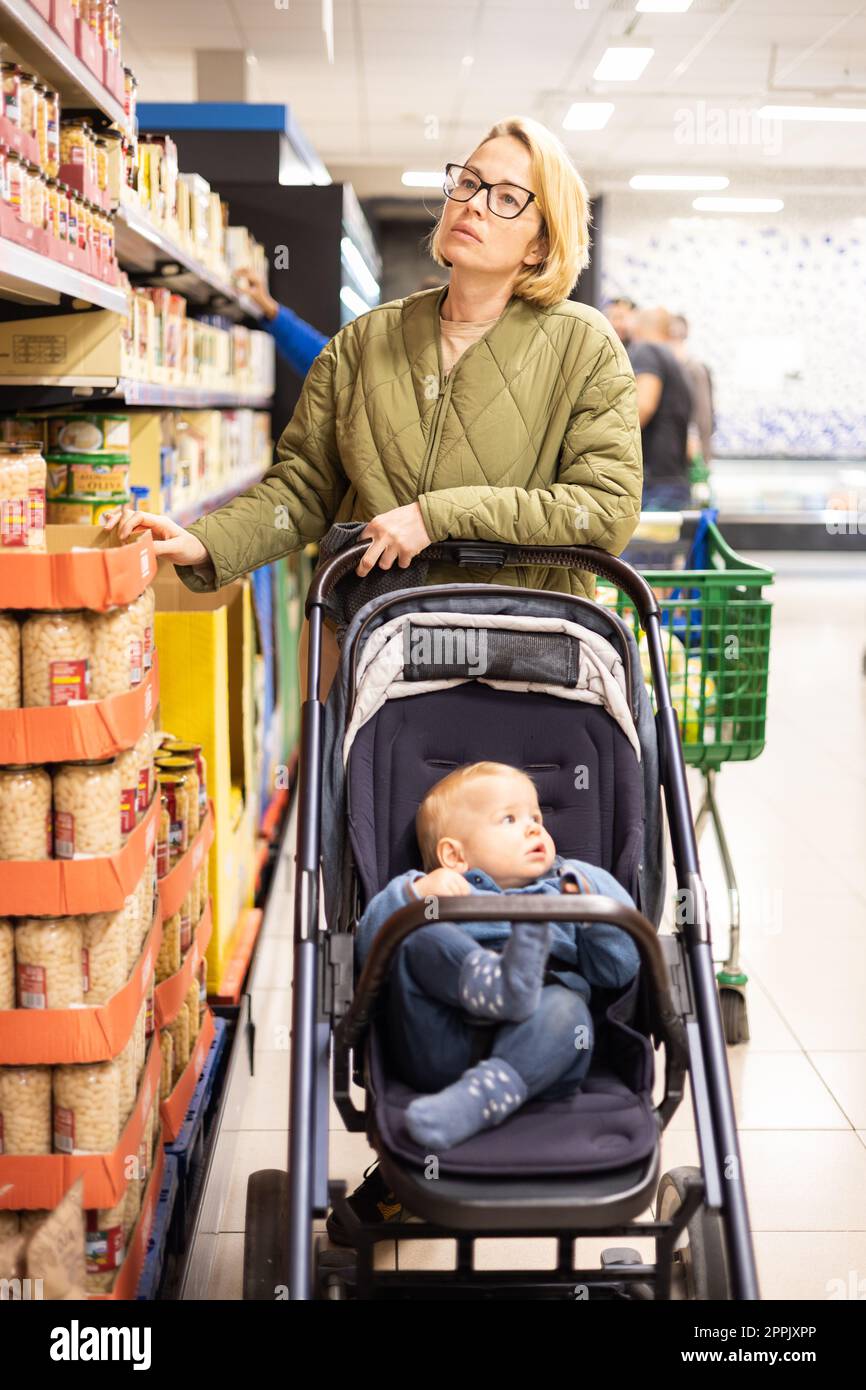 Casualy kleidete die Mutter, die im Supermarkt Konserven auswählte, mit ihrem kleinen Jungen im Kinderwagen. Stockfoto