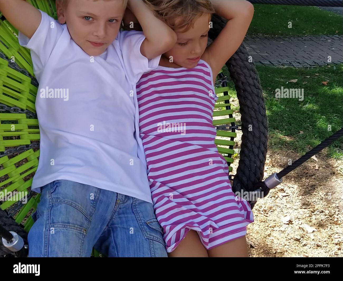 Kinder schwingen auf einer Spinnennetzschaukel. Junge und Mädchen, Bruder und Schwester im Sommer auf dem Spielplatz. Die Schaukel schwingen. Ein 8-jähriger Junge in einem weißen T-Shirt. 6 Jahre altes Mädchen in einem gestreiften rosa Kleid Stockfoto