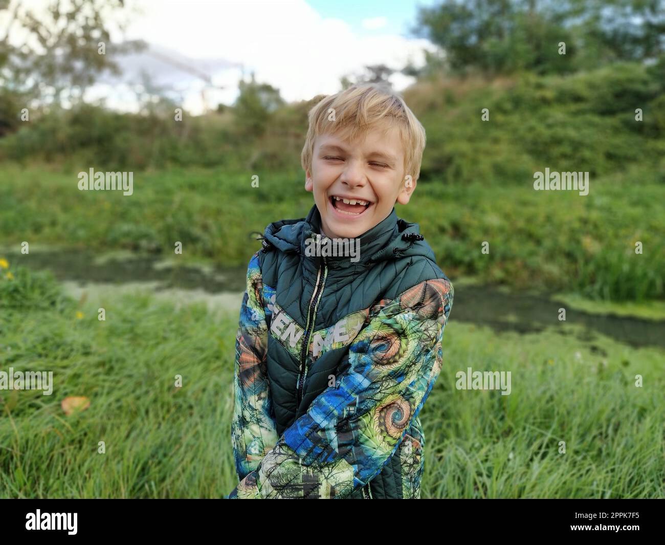 Ein blonder Junge, 8 Jahre alt, lacht im Herbst auf einem Spaziergang an einem Sumpfteich. Ein gewalttätiges Gefühl des Spaßes im Gesicht eines Kindes. Warme Kleidung für jeden Tag. Grüne Jacke. Stockfoto