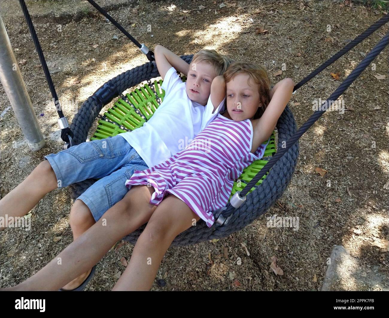 Kinder schwingen auf einer Spinnennetzschaukel. Junge und Mädchen, Bruder und Schwester im Sommer auf dem Spielplatz. Die Schaukel schwingen. Ein 8-jähriger Junge in einem weißen Hemd. 6 Jahre altes Mädchen in einem gestreiften rosa Kleid Stockfoto