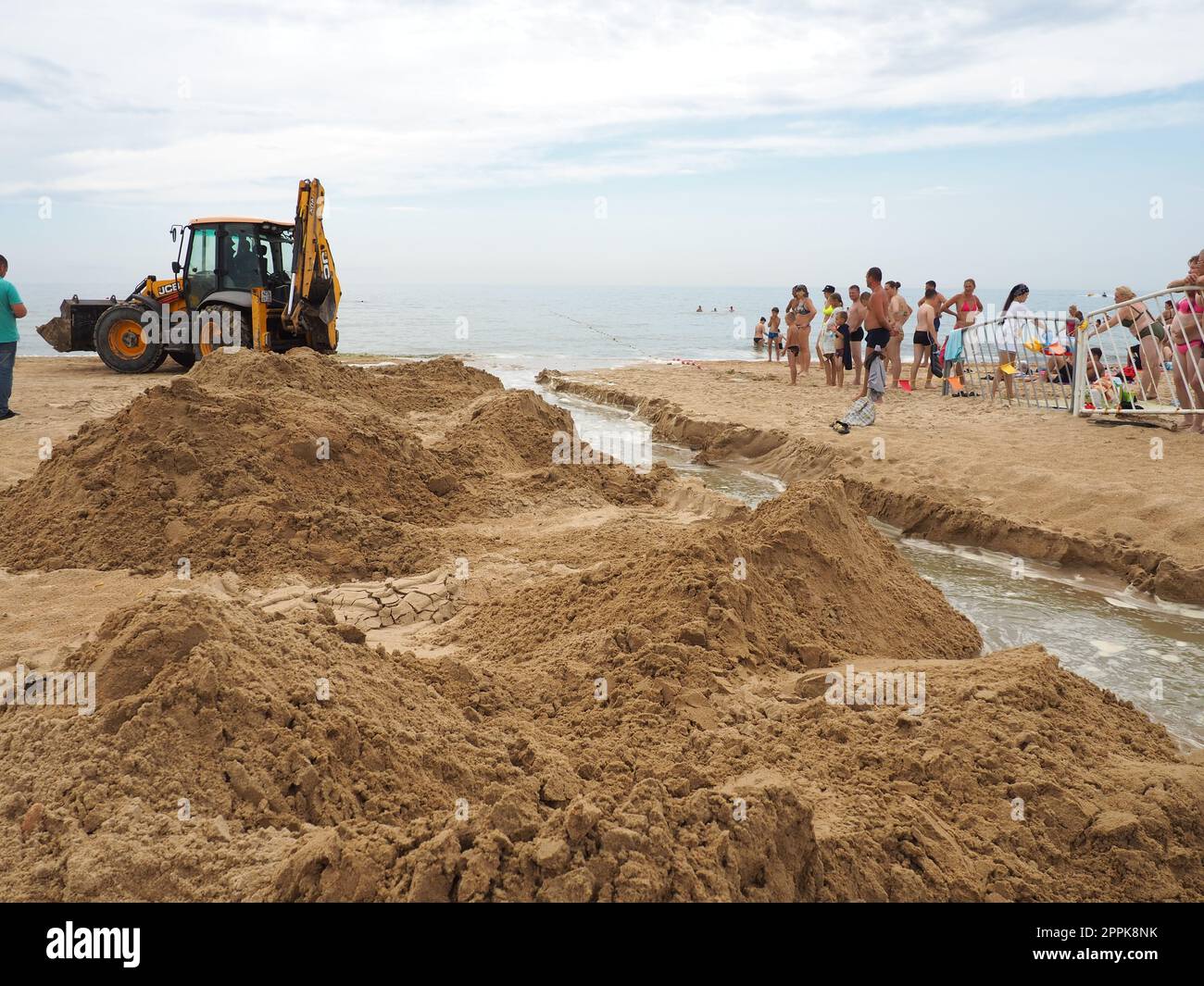 Anapa, Russland, 19. August 2021 Ein Traktor gräbt einen Graben an einem Sandstrand, um verschüttetes Abwasser nach langen Regenfällen abzuleiten. Leute, die Touristen sehen sich Erdbauarbeiten an. Abstieg des Wassers ins Meer Stockfoto