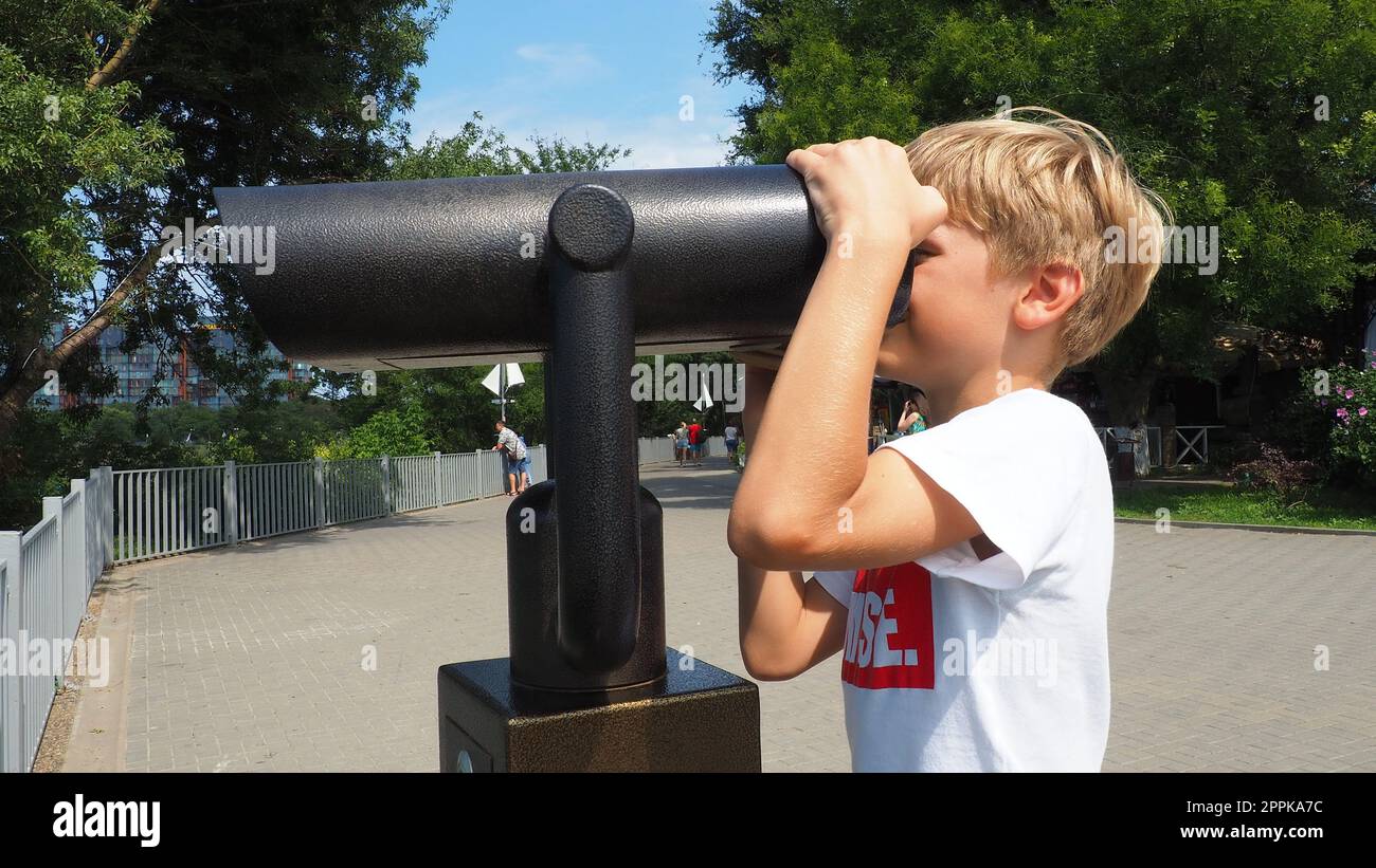 Anapa, Russland, 23. August 2021 Ein Junge schaut durch ein Teleskop. Ein 9-jähriges kaukasisches Kind hält ein großes marines Teleskop mit seinen Händen. Die Straße der Resortstadt und Wandertouristen Stockfoto