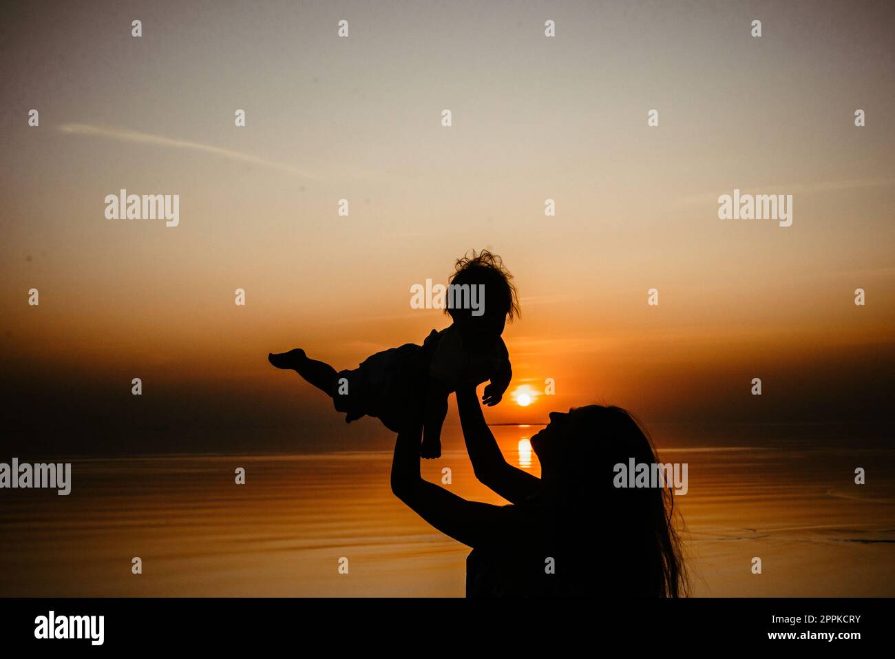 Mama spielt mit dem Kind, lacht zusammen, kreist in ihren Armen und wirft auf. Vor dem Hintergrund des Meeres und des Himmels. Silhouette Stockfoto