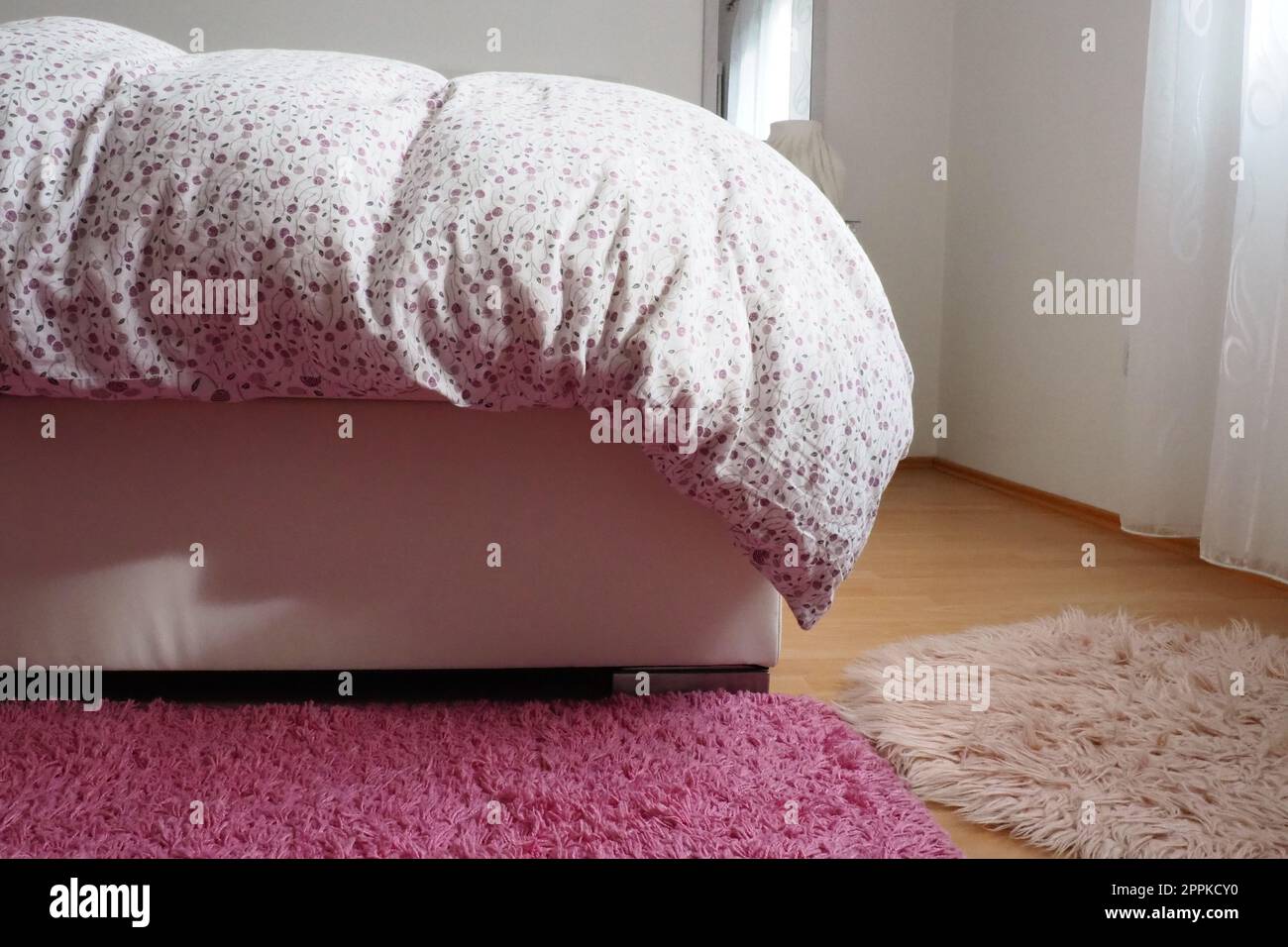 Gemütliches kompaktes Schlafzimmer, weiße Möbel, rosafarbener Teppich und Tulle-Vorhänge. Flauschige Federdecke über das Doppelbett geworfen. Romantischer Morgen. Licht aus dem Fenster. Die Decke hängt bis zum Boden. Stockfoto