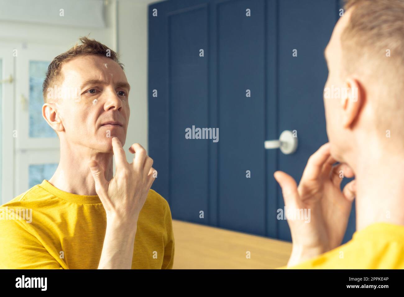 Männer mittleren Alters feuchtigkeitsspendende Creme auf klare Gesichtsbereiche auftragen. Männliches Porträt im Spiegel. Hautpflege-Morgenroutine. Stockfoto