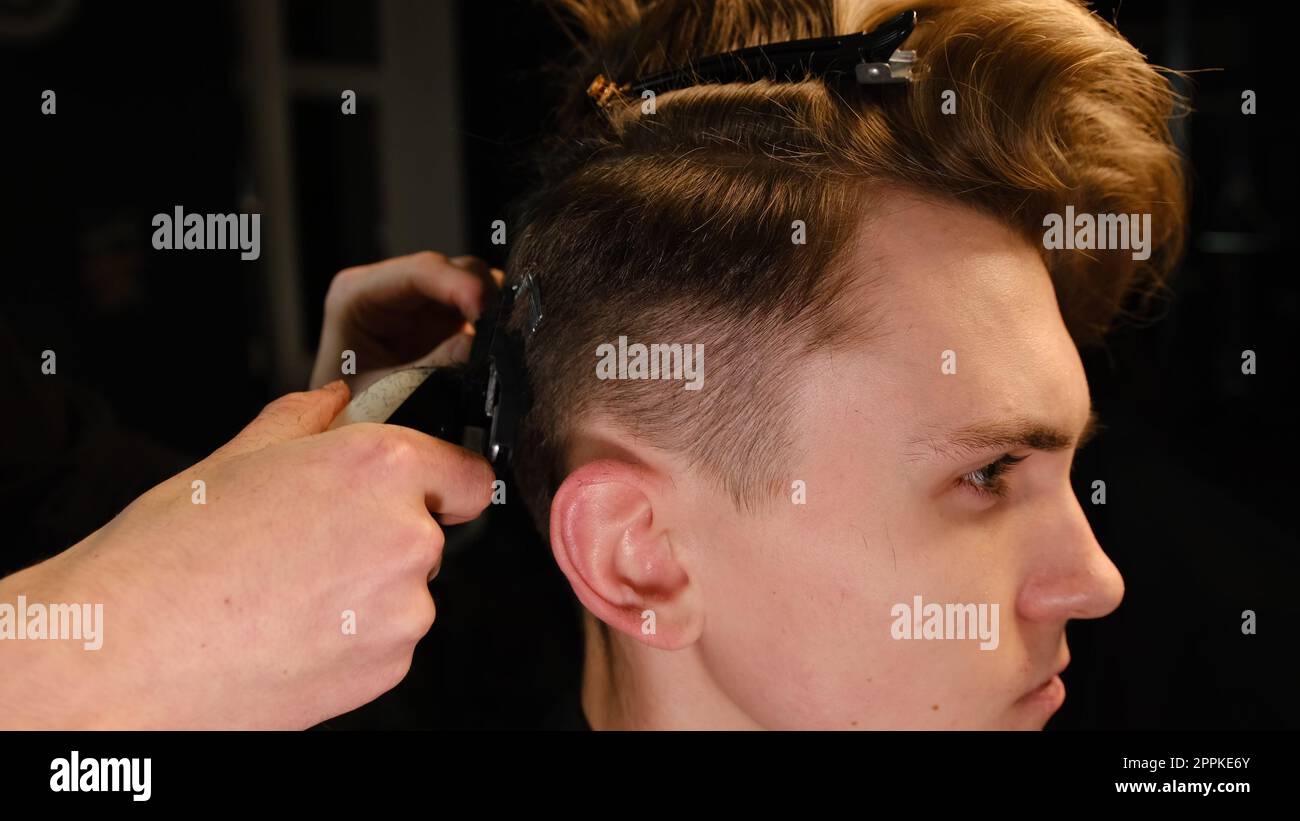 Friseur und Haarschneider für Herren mit Haarschneider in einem Friseur oder Friseursalon. Friseurservice in einem modernen Friseursalon mit dunklem Licht und warmem Seitenblick Stockfoto