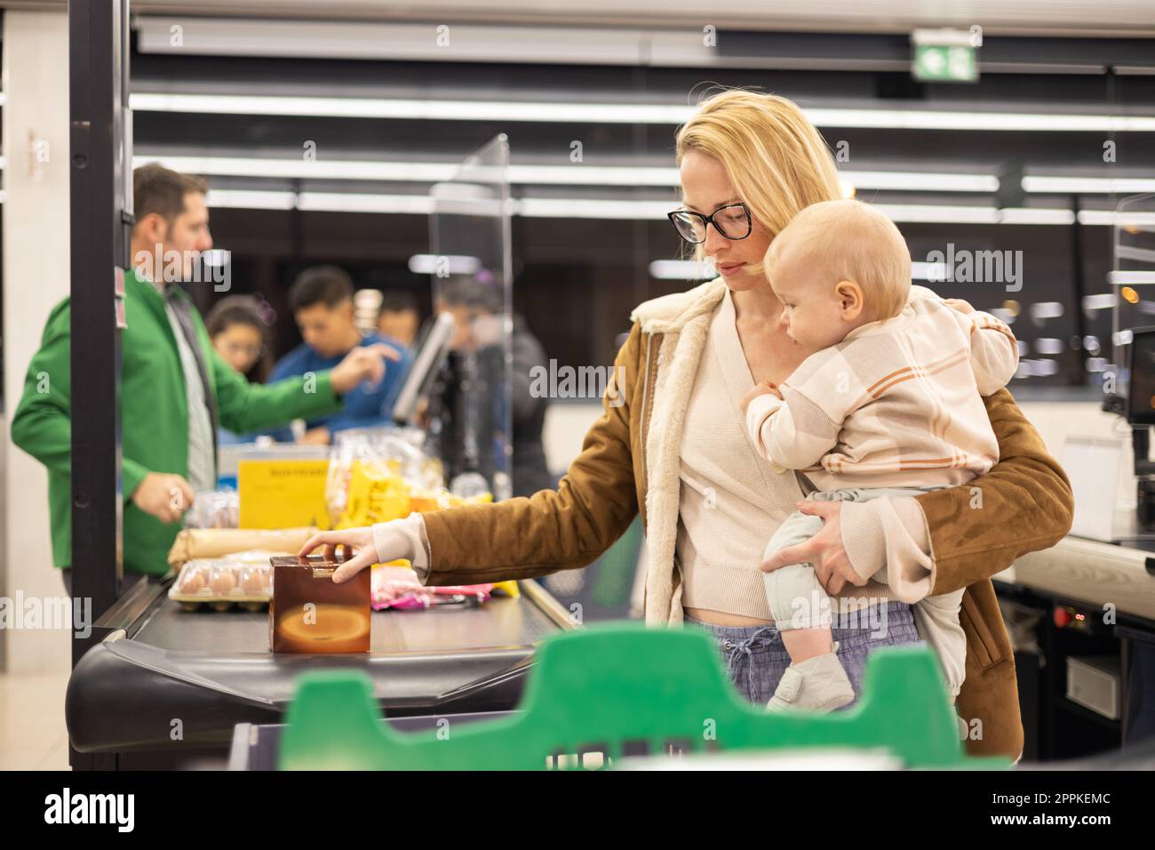 Mutter kauft mit ihrem kleinen Jungen ein und hält das Kind, während sie Produkte an der Kasse im Supermarkt stapelt. Stockfoto