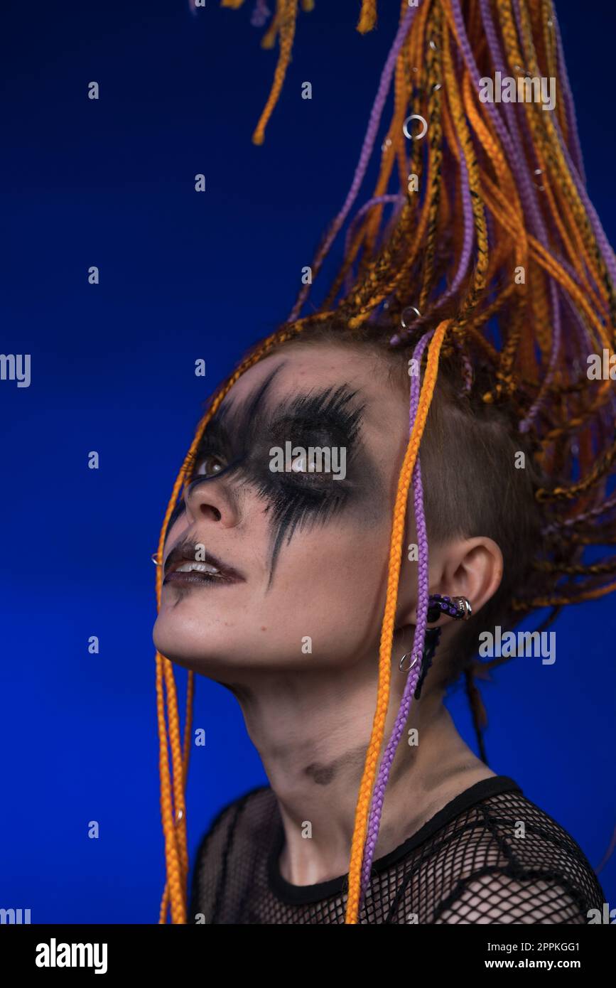 Kinoporträt einer jungen Frau mit grauenschwarzem Bühnenschminke auf dem Gesicht und orangefarbenem Dreadlocks-Haarstil. Studio hat einen blauen Hintergrund aufgenommen Stockfoto