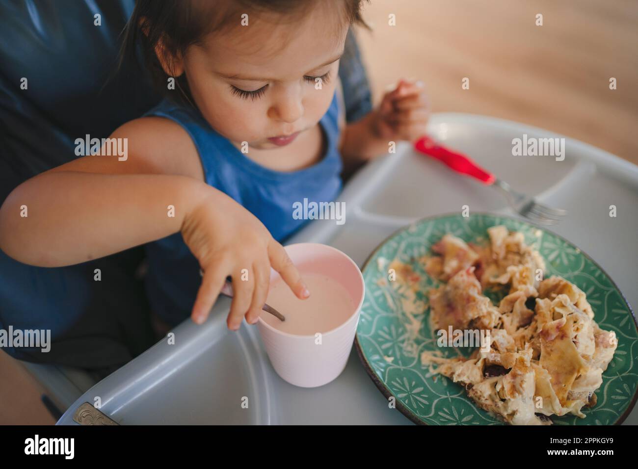 Ein kleines Mädchen lernt, sich selbst zu essen, während es in einem  Kinderstuhl sitzt und mit den Händen und einer Gabel vom Teller isst.  Gesunde Ernährung, Lebensstil Stockfotografie - Alamy