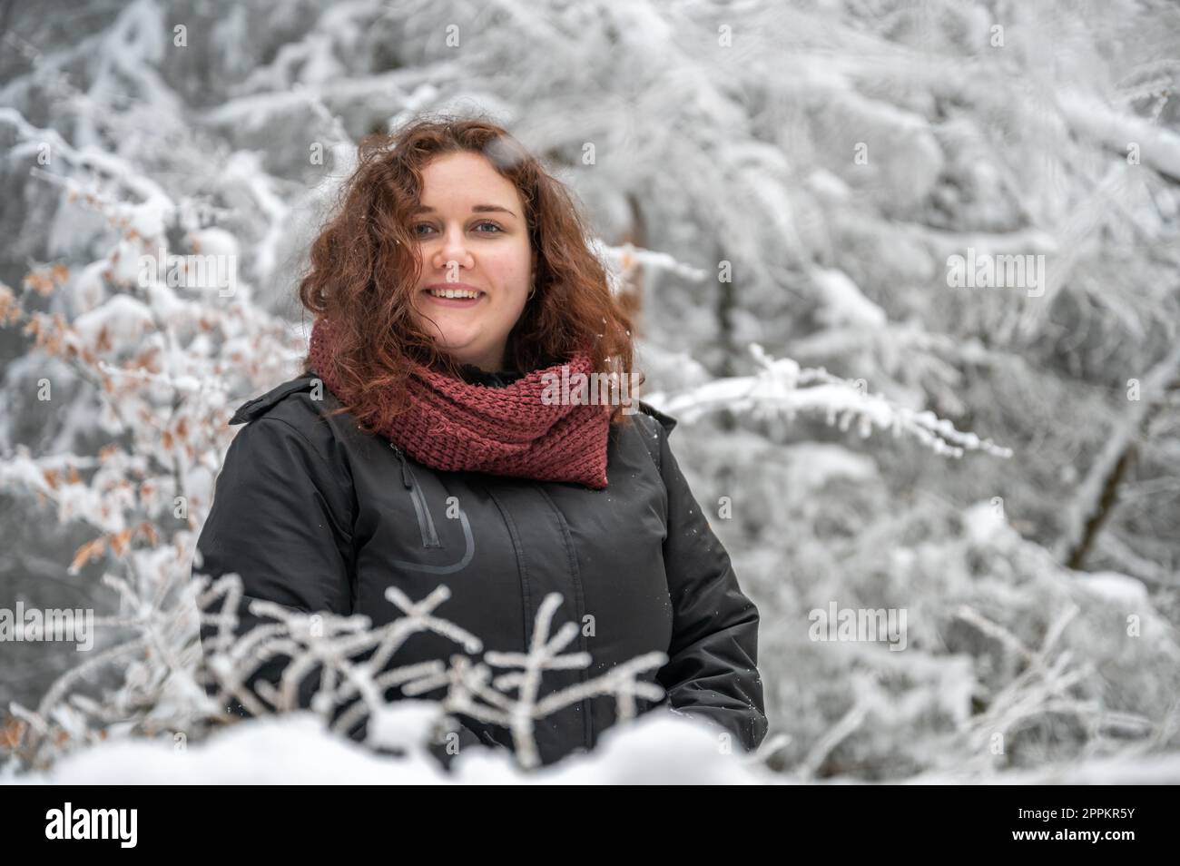 Eine junge Frau mit braunem lockigem Haar lächelt und schaut in die Kamera, während sie im Winter mitten in einem verschneiten Wald steht Stockfoto