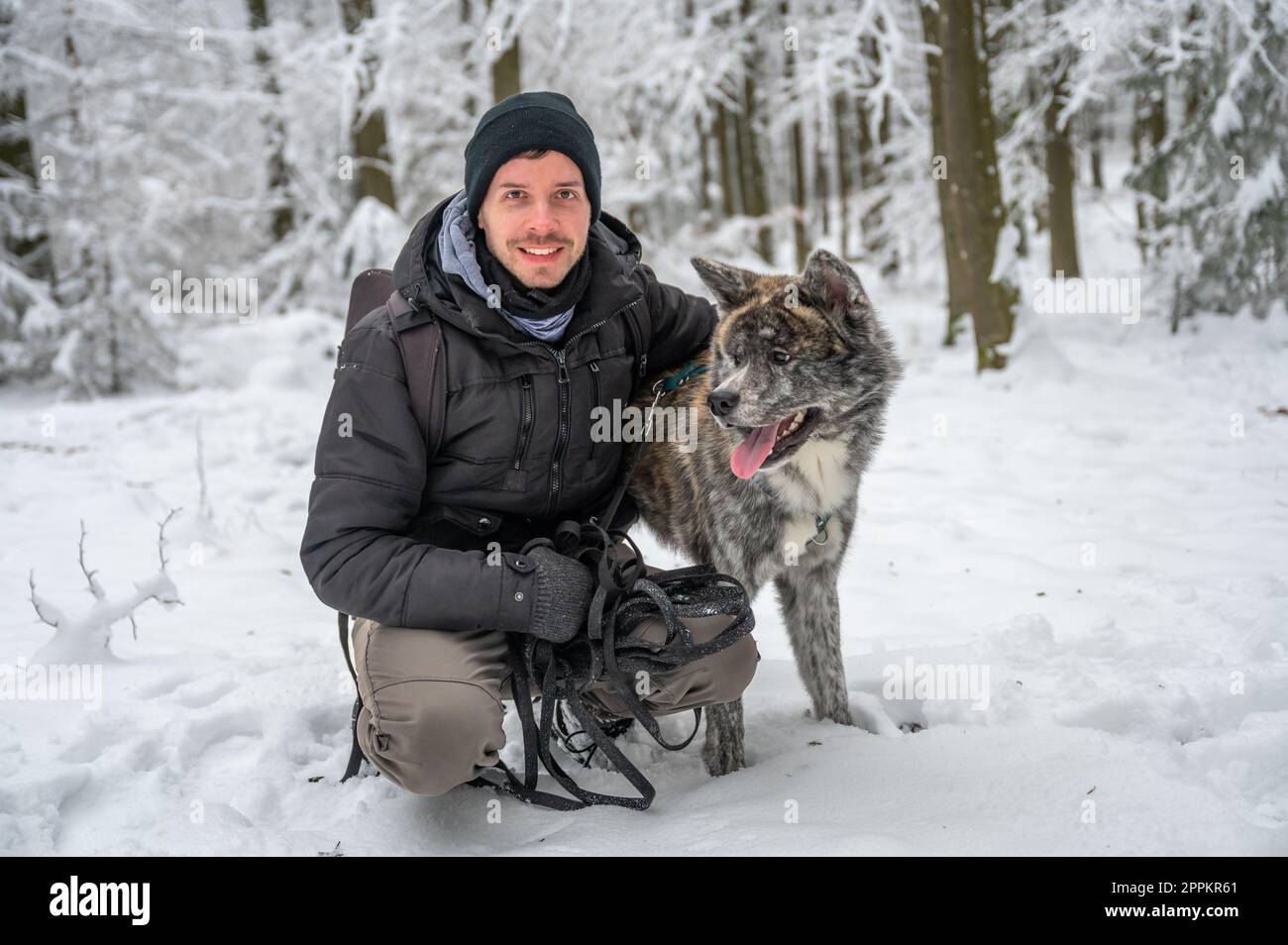 Meister mit seinem grauen Akita Inu Hund im Wald im Winter mit viel Schnee, der Mensch lächelt und schaut in die Kamera, während er seinen Hund umarmt Stockfoto