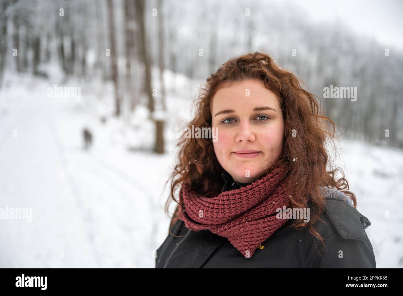 Eine junge Frau mit braunem lockigem Haar und warmer Kleidung lächelt und schaut in die Kamera, geht im Winter mit viel Schnee mit ihrem grauen akita inu Hund im Wald spazieren Stockfoto