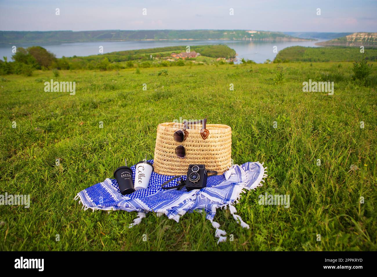 Eine sehr schöne Aussicht auf den See. Ein Strohkorb steht auf einer blauen Decke auf grünem Gras zusammen mit Gläsern, einer Kamera und Kaffeetassen. Erholung im Freien. Stockfoto