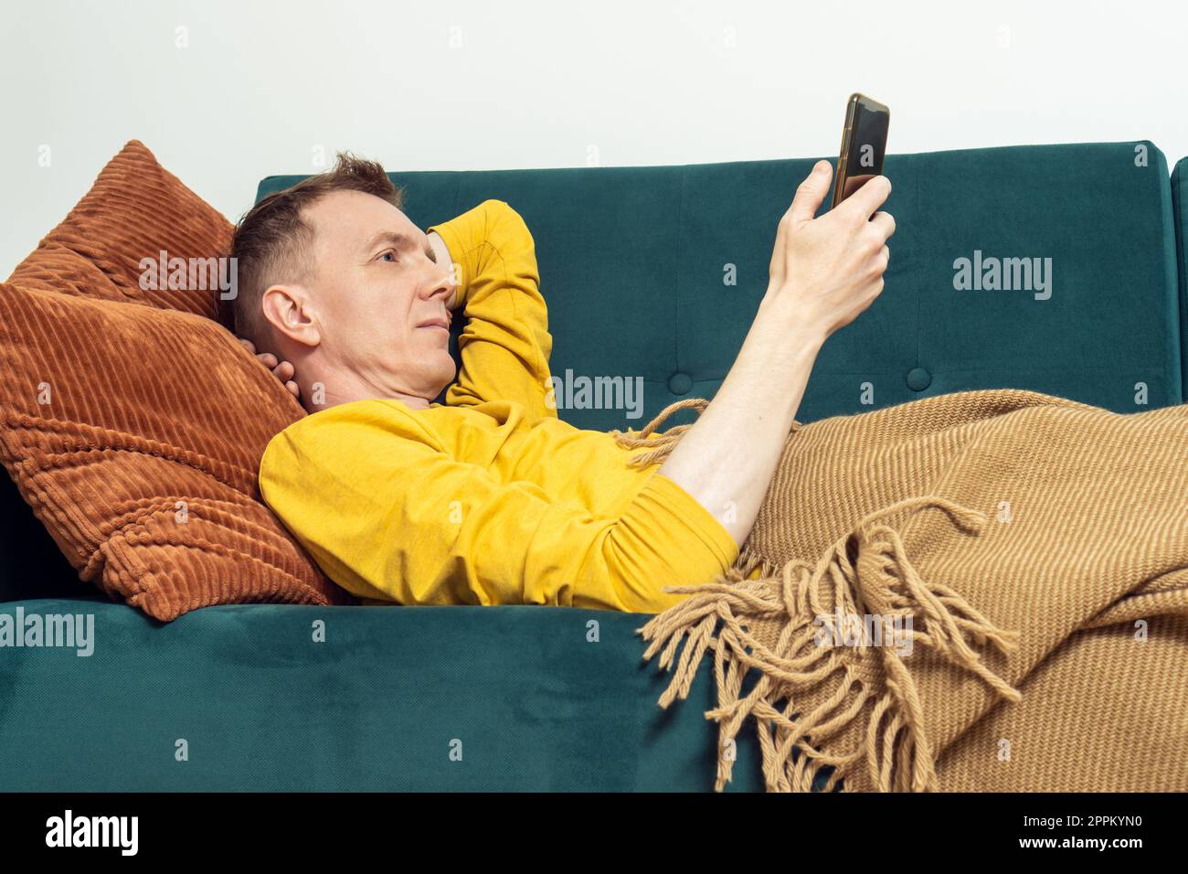 Erwachsener auf dem Sofa, Video auf Smartphone anschauen, Seitenansicht. Person, die sich auf dem Kissen unter der Decke ruht und sich entspannen kann. Stockfoto